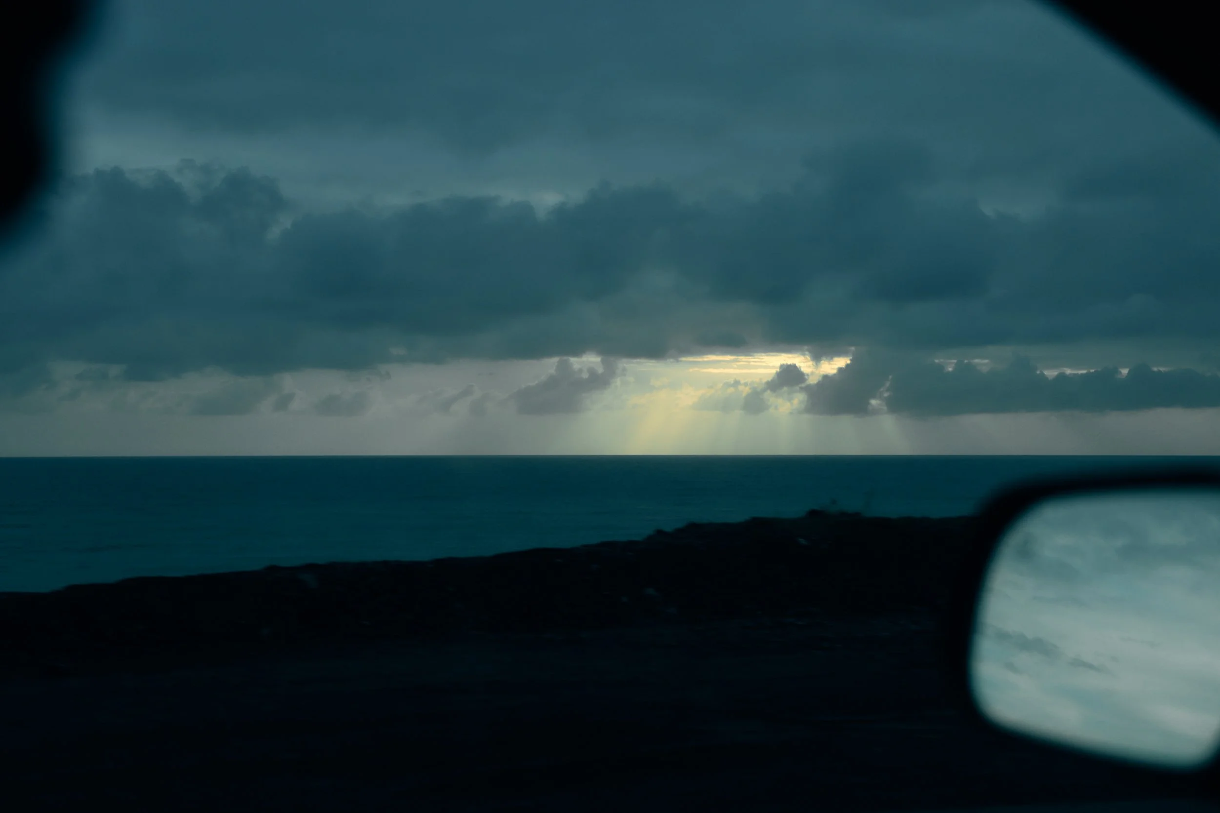 View of a stormy sky over the ocean as seen from a moving vehicle, with part of the car's side mirror visible in the foreground.