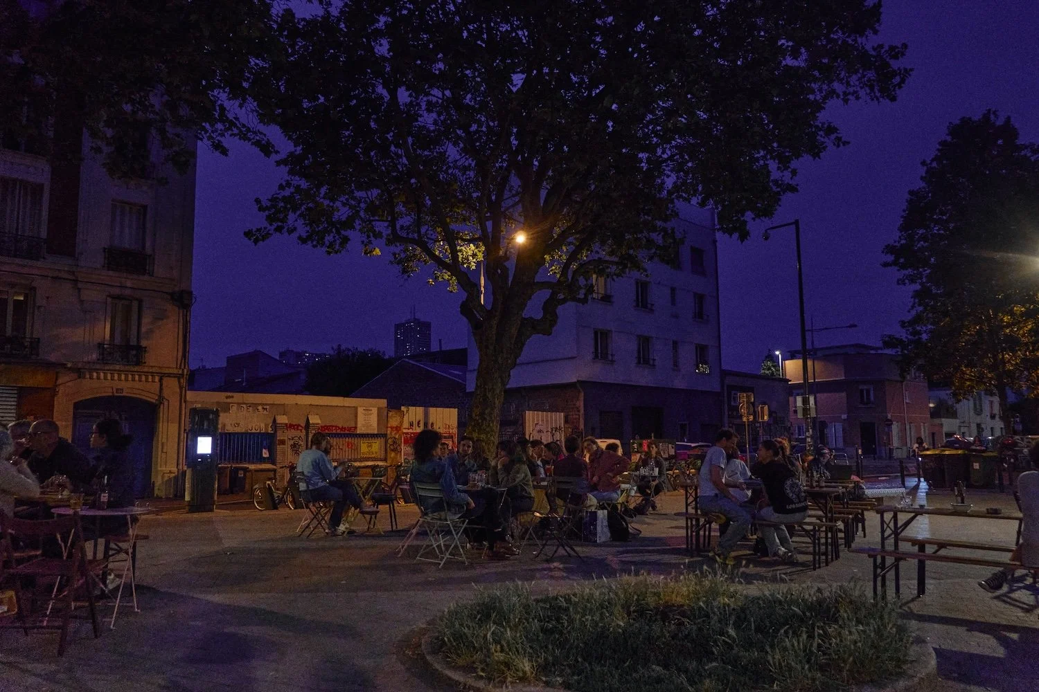 People sitting and socializing at outdoor tables in a city square at night, illuminated by streetlights and a large tree in the center.