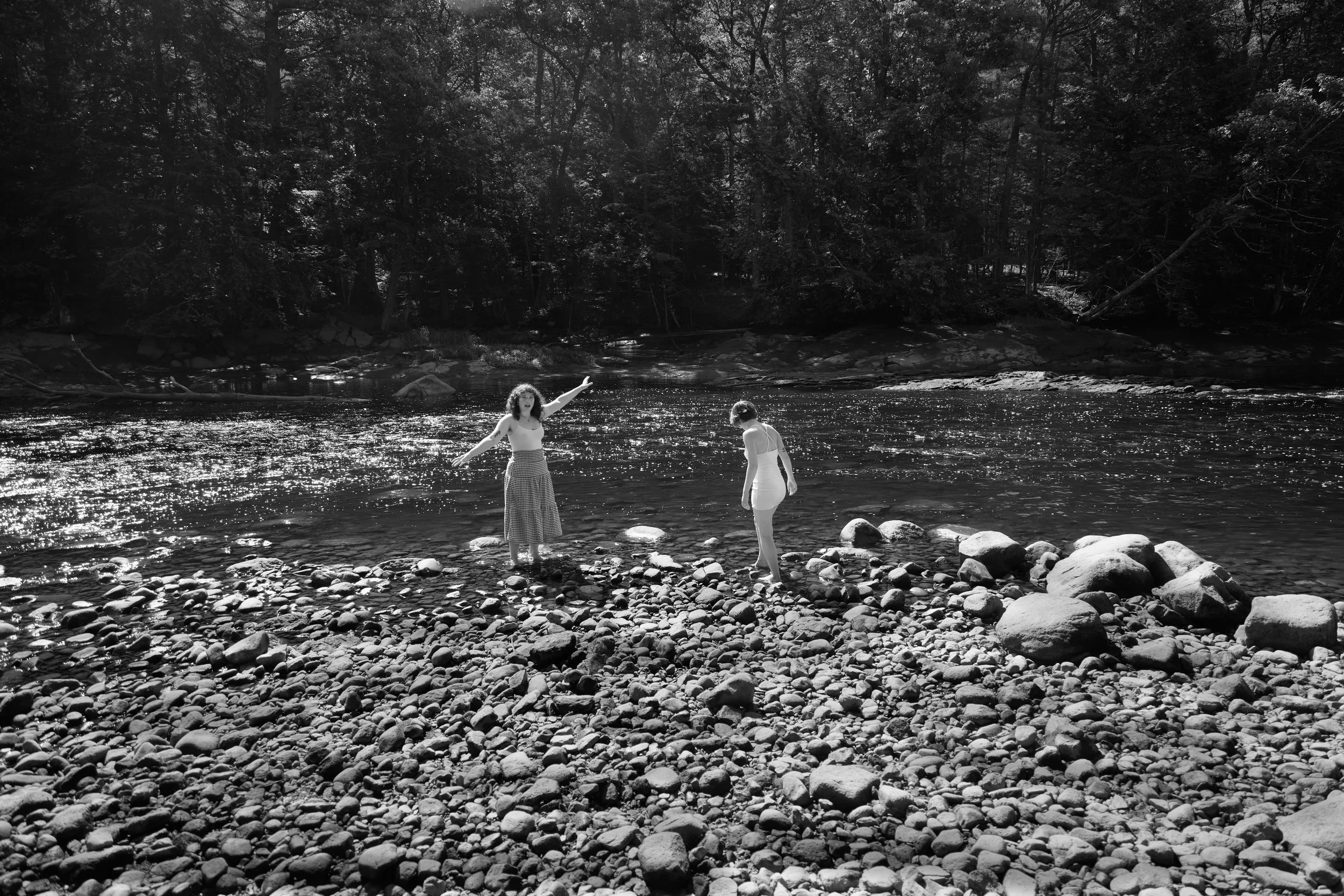 Two women are walking in a river surrounded by a rocky shore and dense trees in the background.