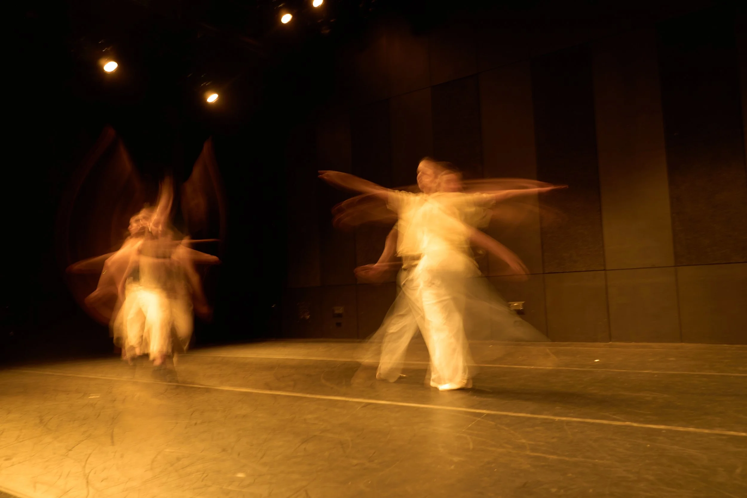 A stage performance of two dancers in white dresses spinning, captured with motion blur in a dimly lit theater.