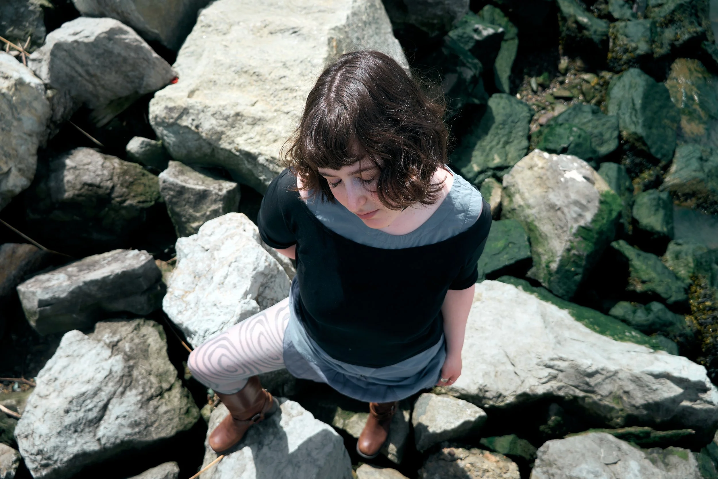 A woman with curly brown hair stands on rocks near a stream or river, looking down with her eyes closed, surrounded by greenery.