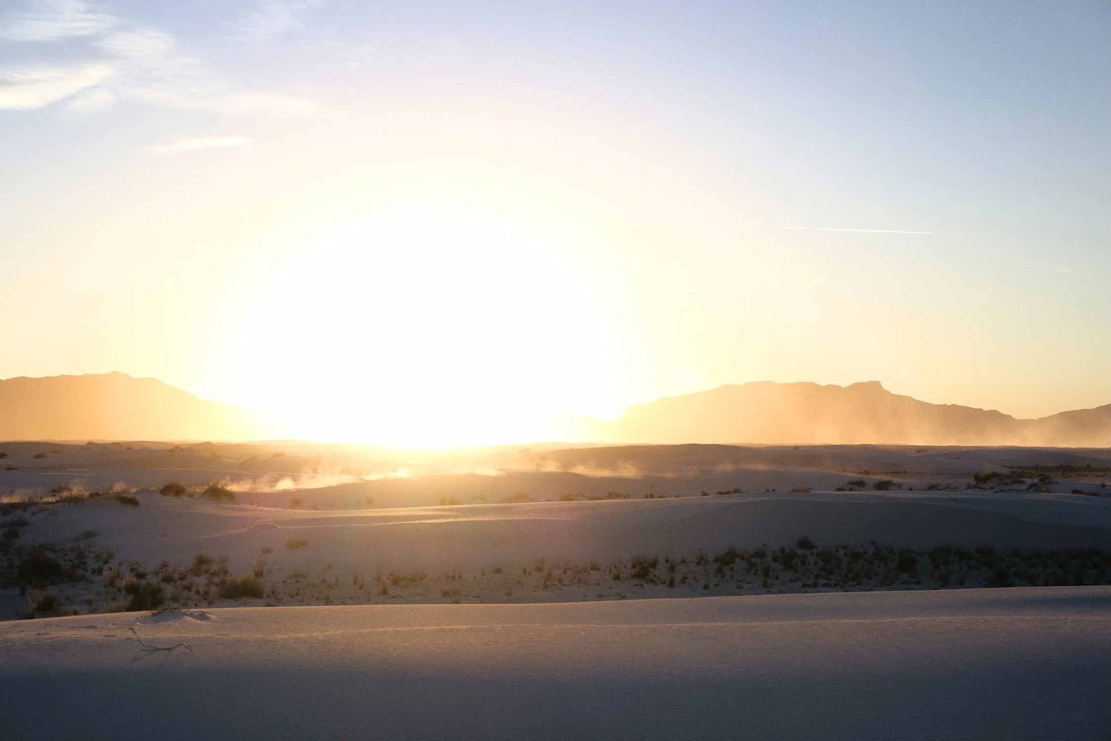 A desert landscape at sunset with mountains in the distance and a bright, glowing sun near the horizon. The sky is mostly clear with a few clouds.