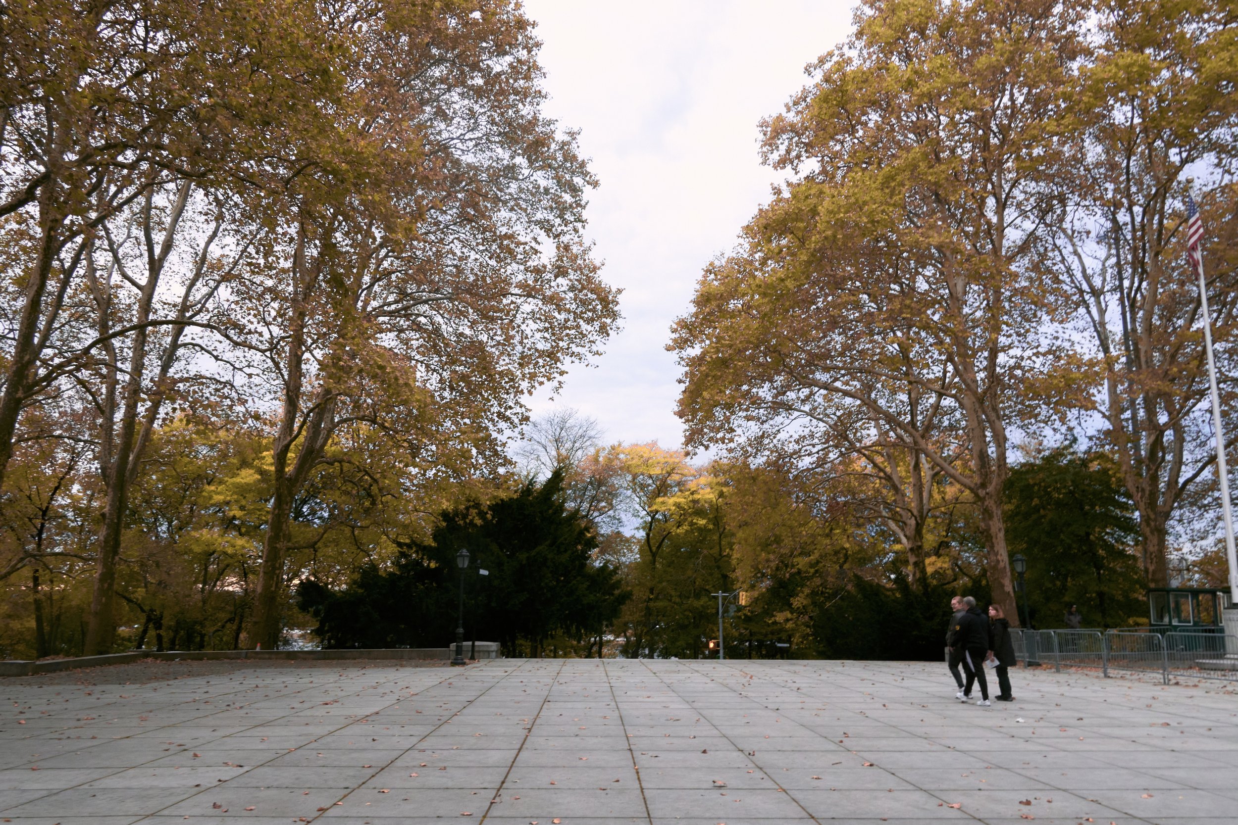 Park with large trees with autumn foliage, an American flag on a flagpole, and a group of three people walking on a paved open space.
