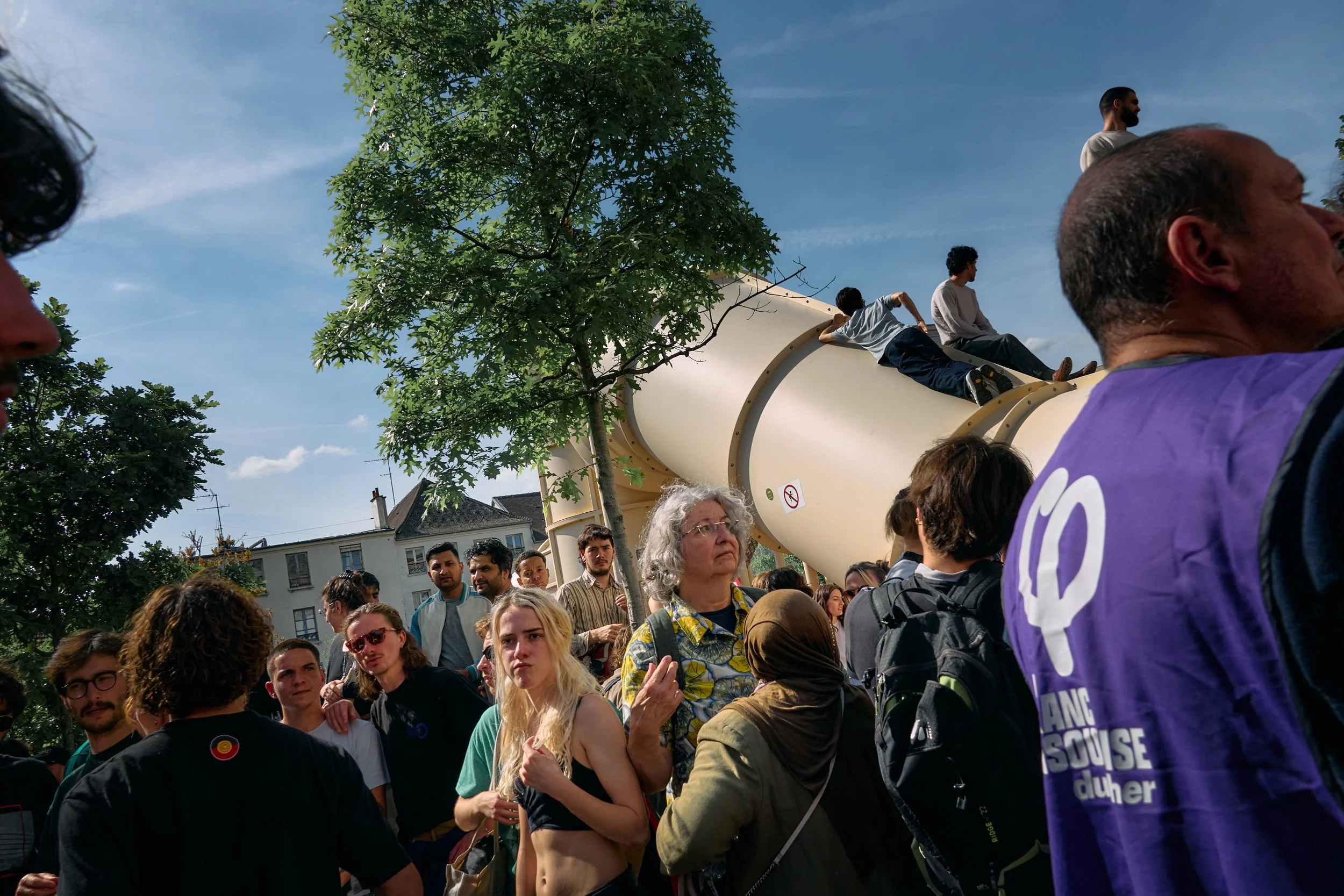 A large crowd of diverse people gathered outdoors under a partly cloudy sky, standing near a large white slide with a warning sign, with some individuals sitting on or climbing the slide, and others watching.