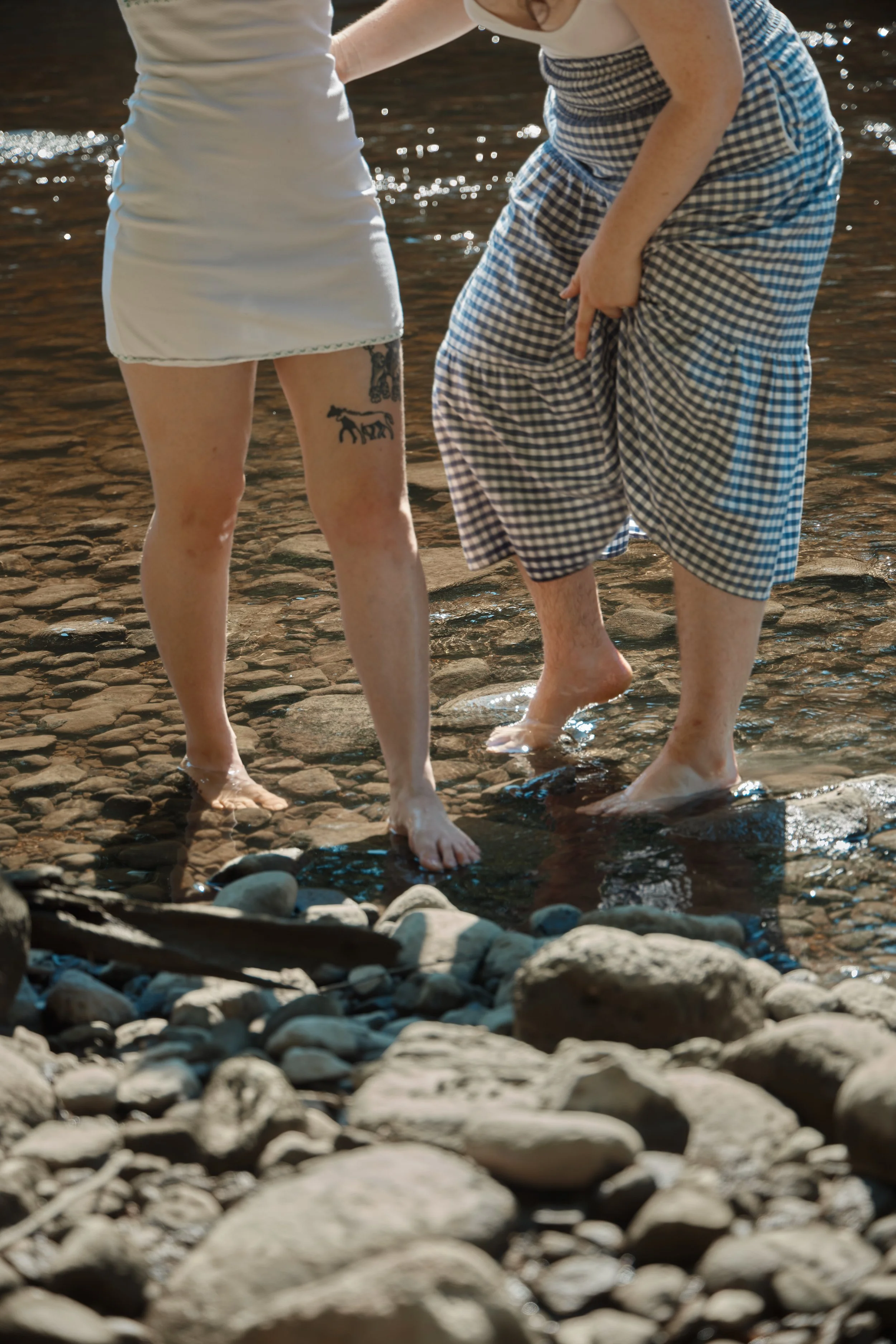 Two people standing in a shallow rocky river, only their legs and lower torsos visible, with one person pointing downward at the rocks.