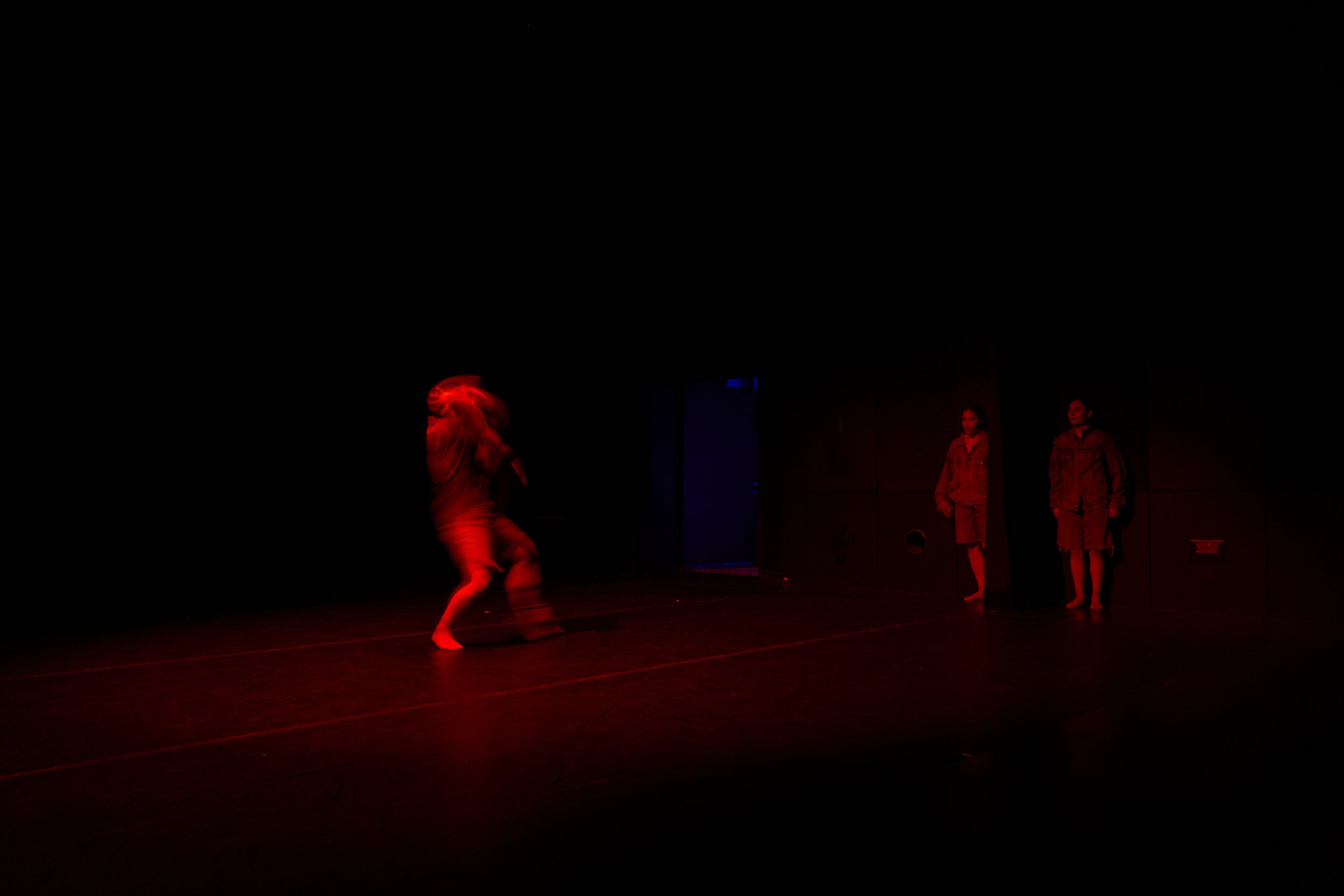 A dance performance on stage with dark background, illuminated by red and blue lighting. Two dancers are visible, one in motion, and two other individuals are standing at the back, observing or preparing to perform.