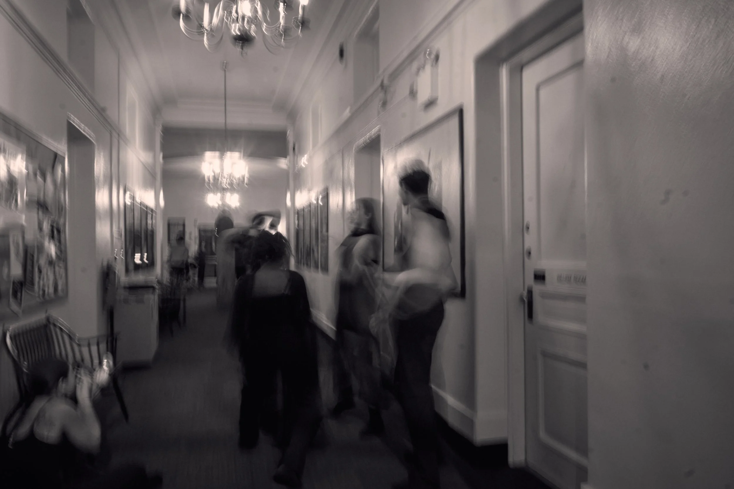A hallway with ornate chandeliers on the ceiling, framed pictures on the walls, and several people standing and talking. There are chairs against the walls and a bulletin board. The image is in black and white.