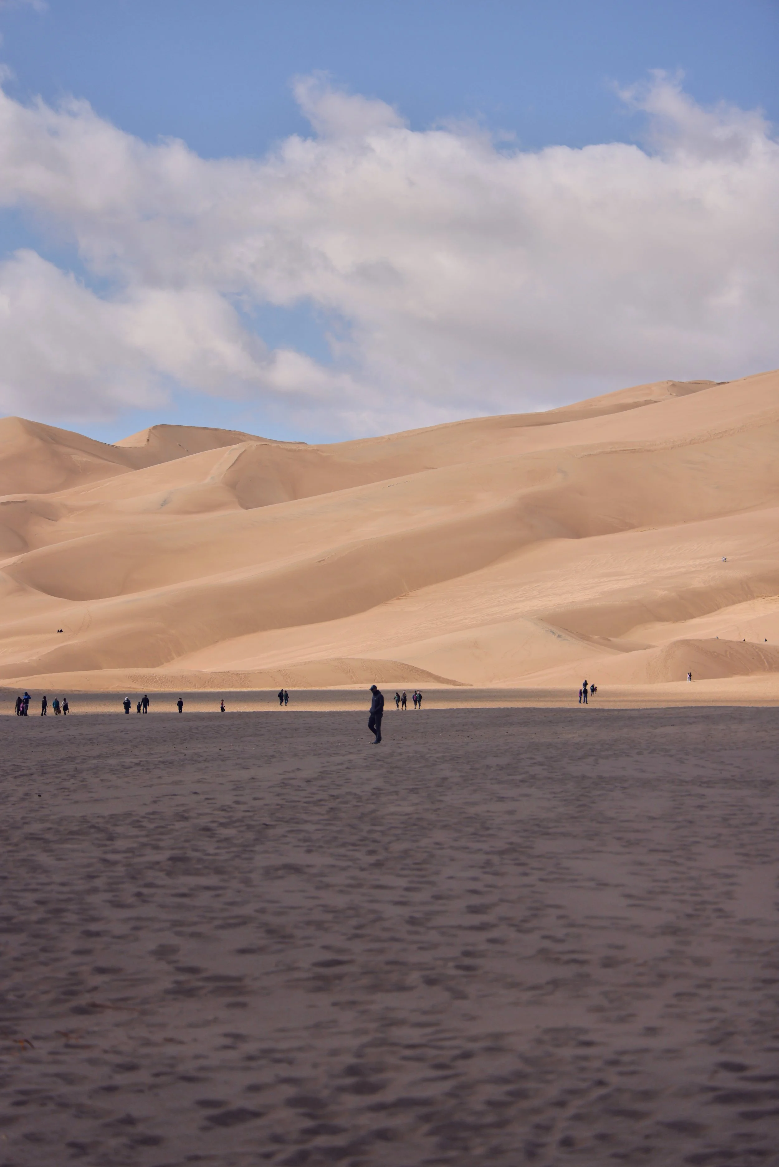 Desert landscape with large sand dunes under a partly cloudy sky, and small silhouettes of people walking on the flat ground in the foreground.