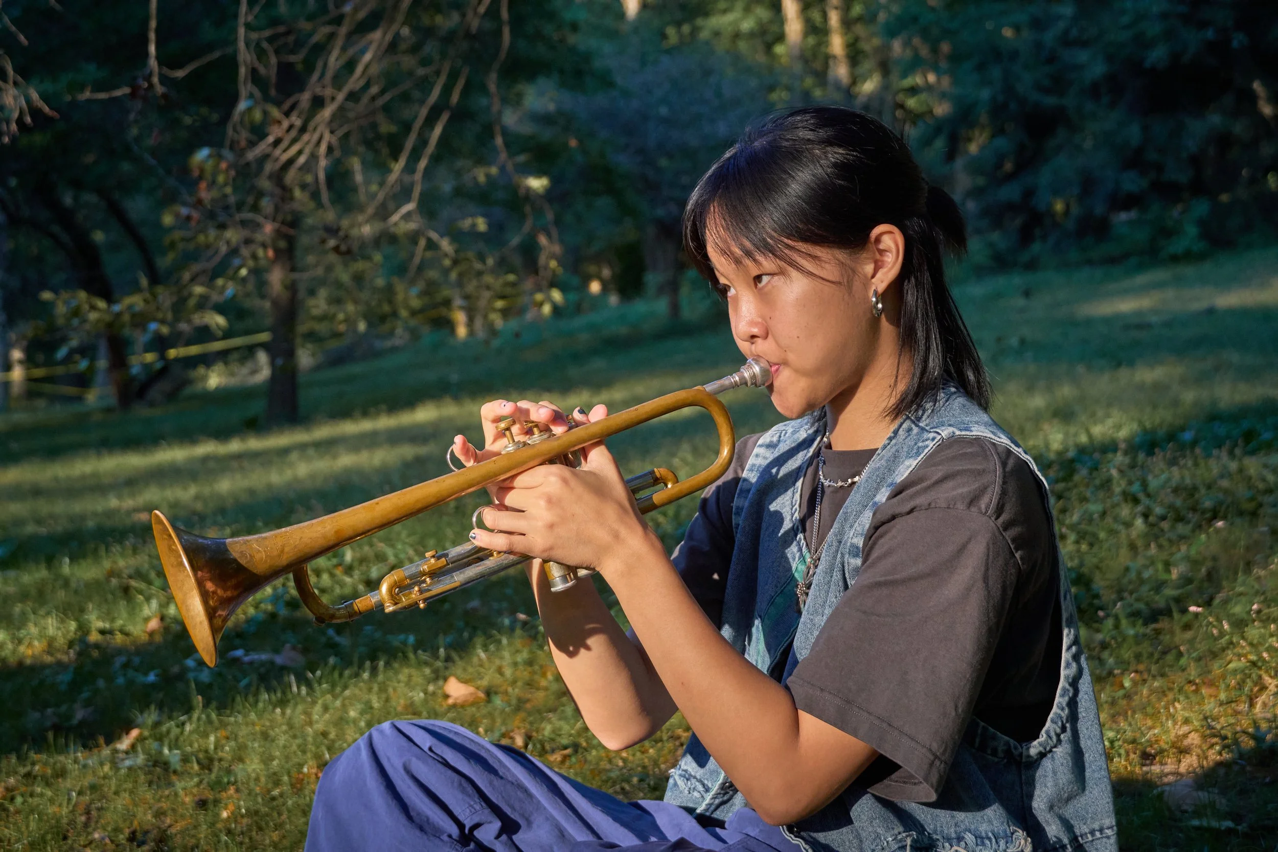 Young woman with dark hair and earrings playing a trumpet outdoors in a park during daytime.