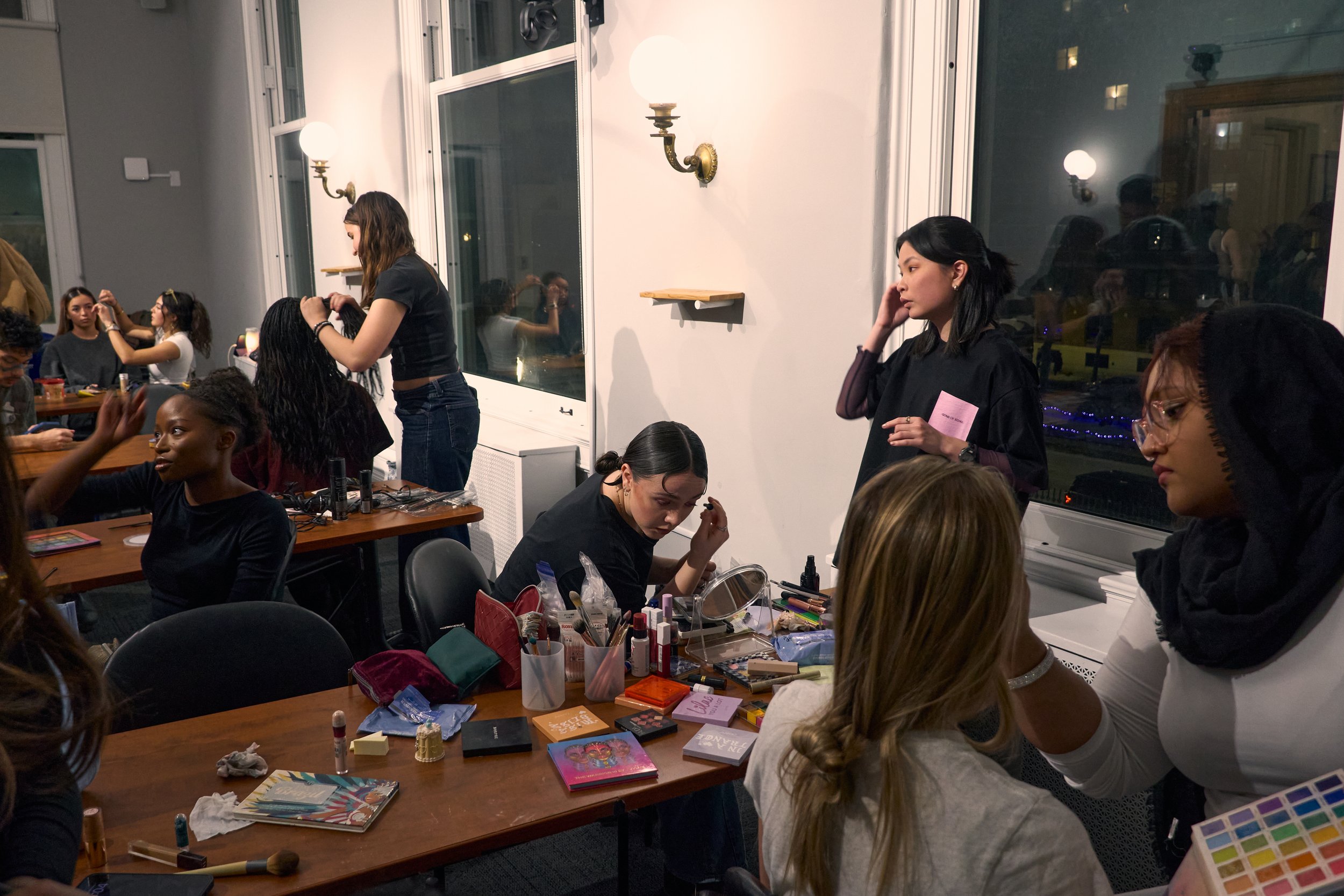 A group of women gathered in a room, sitting at tables with makeup and beauty products, some women applying makeup, and others waiting or grooming. The room has large windows, wall sconces, and a reflection of lights outside.