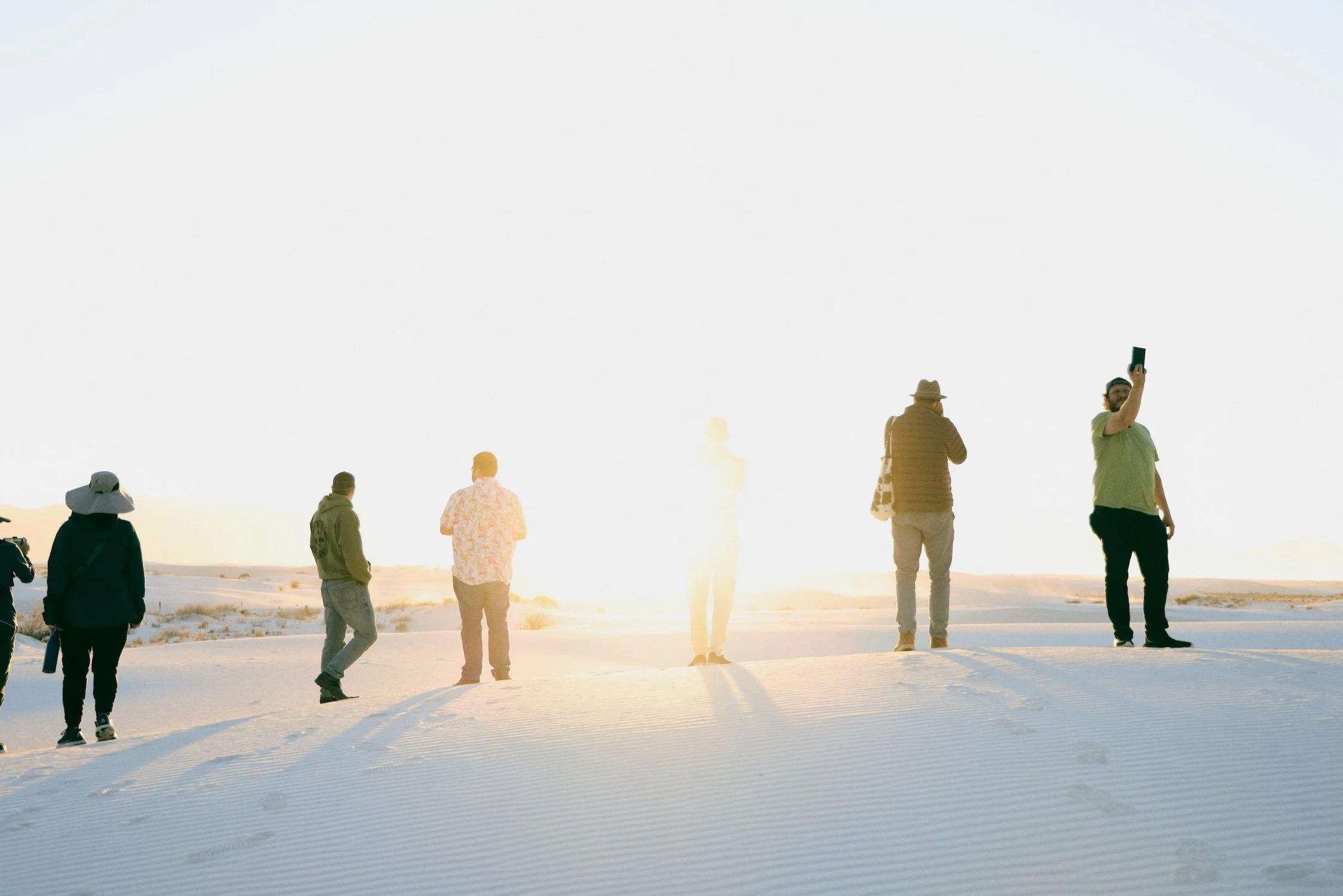 Several people are walking on a snow-covered landscape during sunset, some are taking photos, and all are dressed in warm clothes.