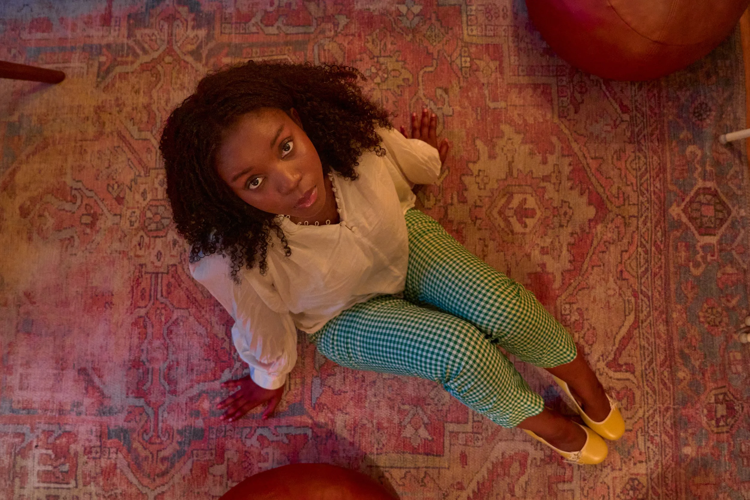 From an overhead perspective, a woman with curly hair is sitting on a patterned rug, looking up at the camera. She is wearing a cream-colored blouse and green checkered pants, with yellow shoes placed on the rug.