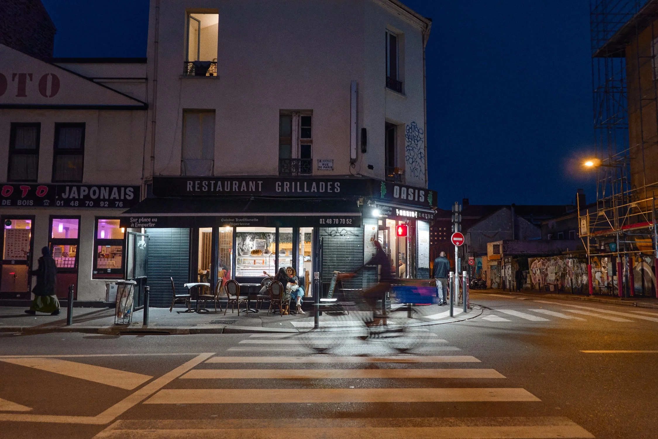 Nighttime street scene in Paris with a restaurant named Oasis, people sitting outside, and a building with lit windows and graffiti on the wall.