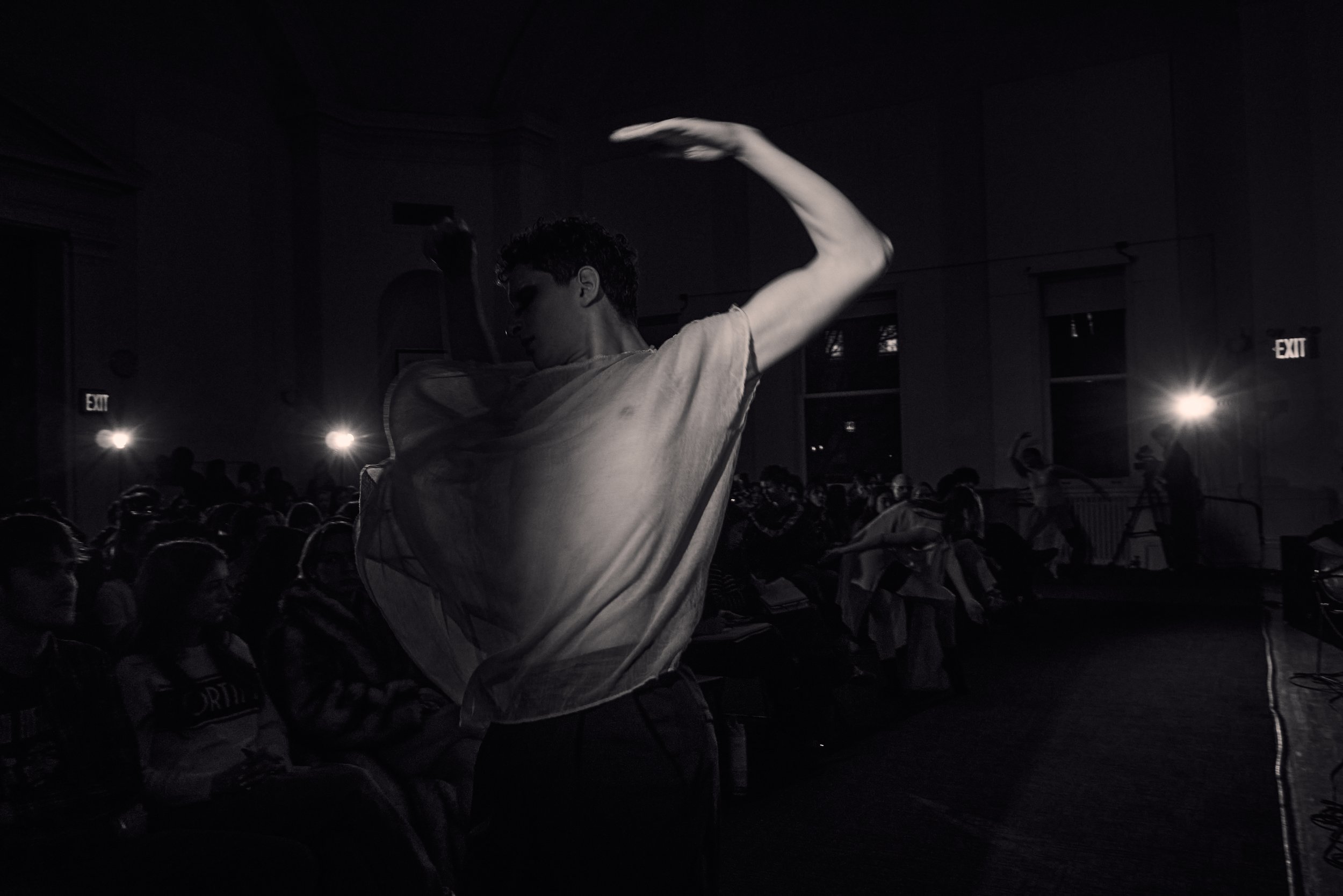 A person with curly hair dancing in a dark room with an audience, illuminated by stage lights and a glow from cellphone screens.