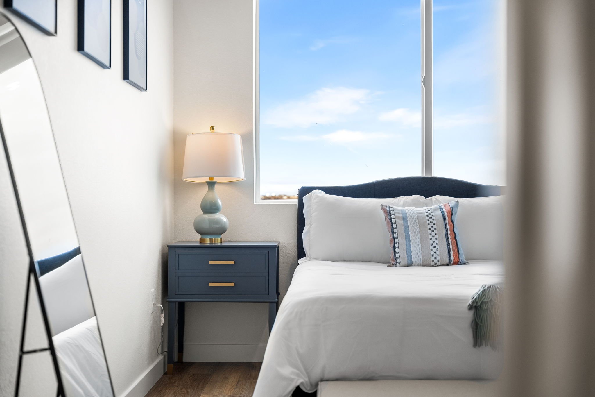 A modern bedroom with a bed, a blue nightstand, a lamp, and a window showing a blue sky.