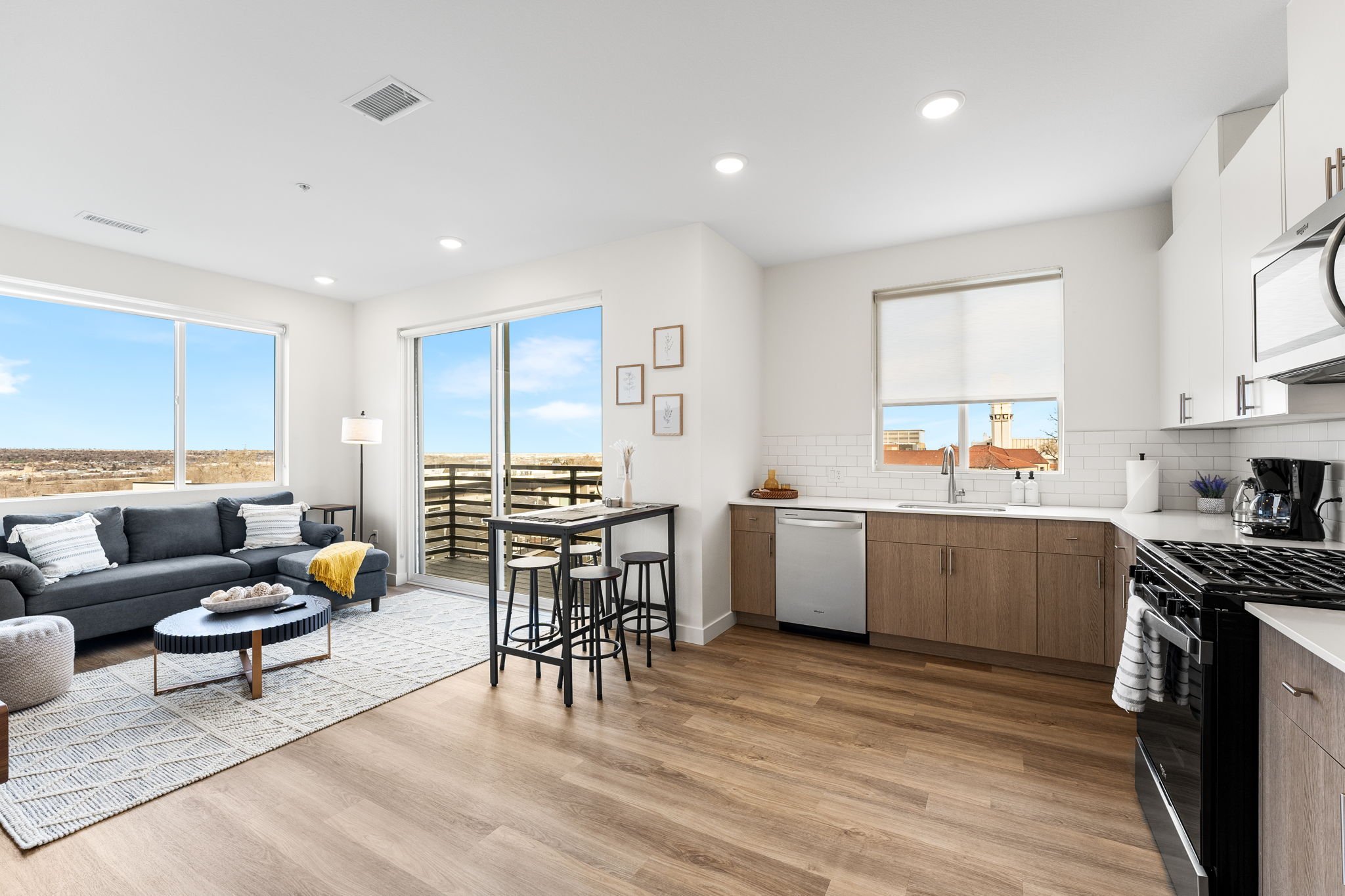 Living room and kitchen with large windows, gray sofa, fireplace, dining table with stools, kitchen cabinets, white countertops, and stainless steel appliances.