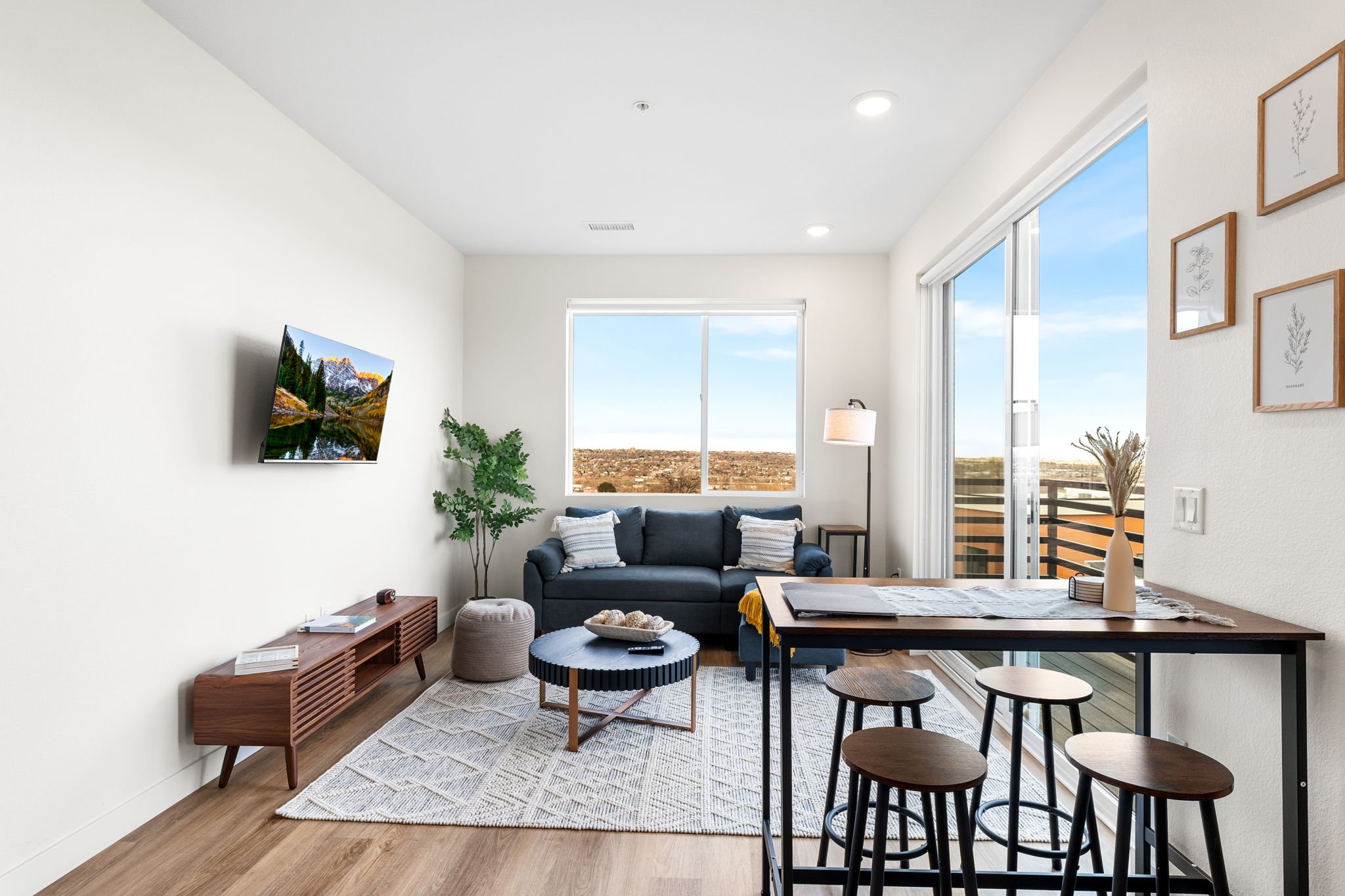 Living room with white walls, hardwood flooring, a dark blue sofa with white and striped pillows, a round coffee table, and a TV mounted on the wall. A large window and sliding glass door lead to a balcony with outdoor views. Decor includes a potted 