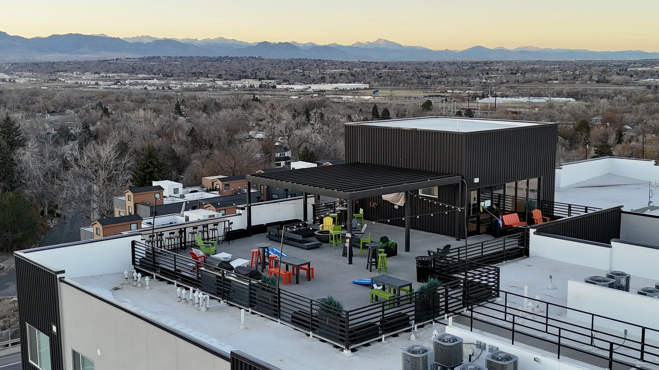 Rooftop terrace with colorful outdoor furniture, string lights, and potted plants, overlooking a suburban area with trees and distant mountains at sunset.