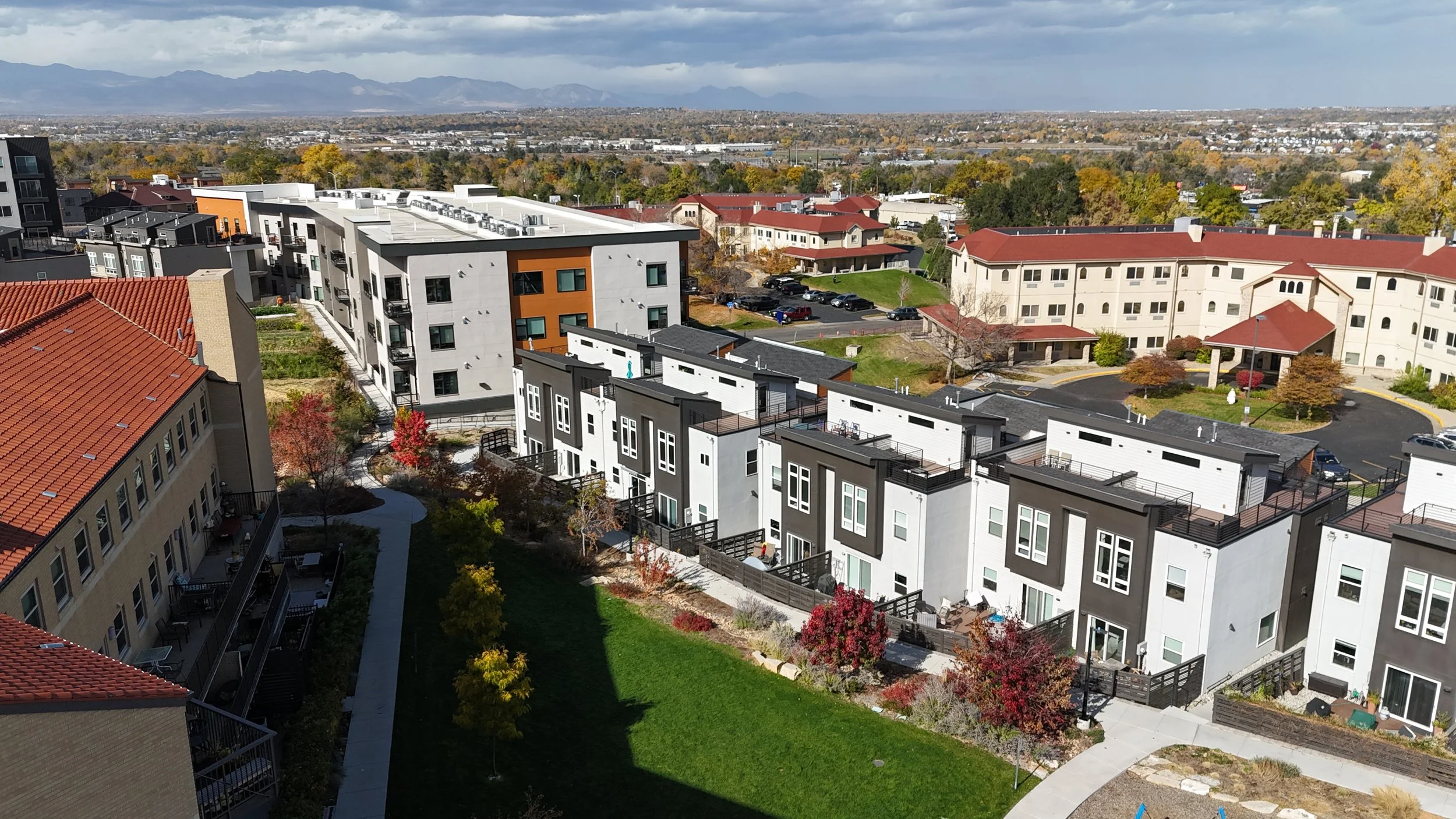 Aerial view of a residential apartment complex with modern and traditional buildings, landscaped gardens, parking lots, and a distant mountain range under a partly cloudy sky.