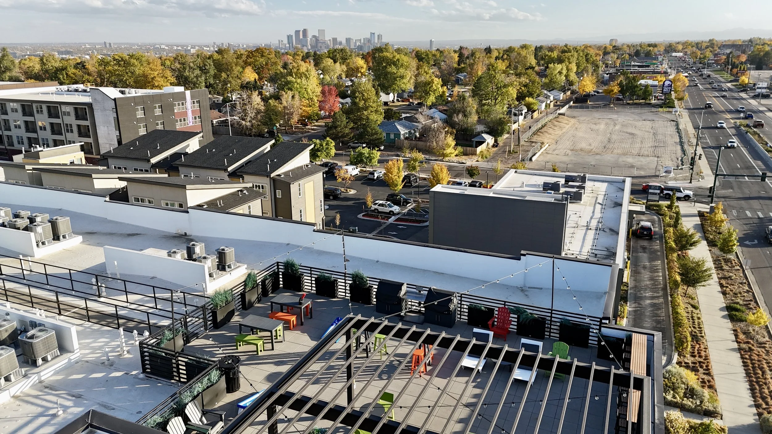 A rooftop terrace with colorful chairs and tables, surrounded by potted plants, overlooking a cityscape with buildings, trees, and streets.