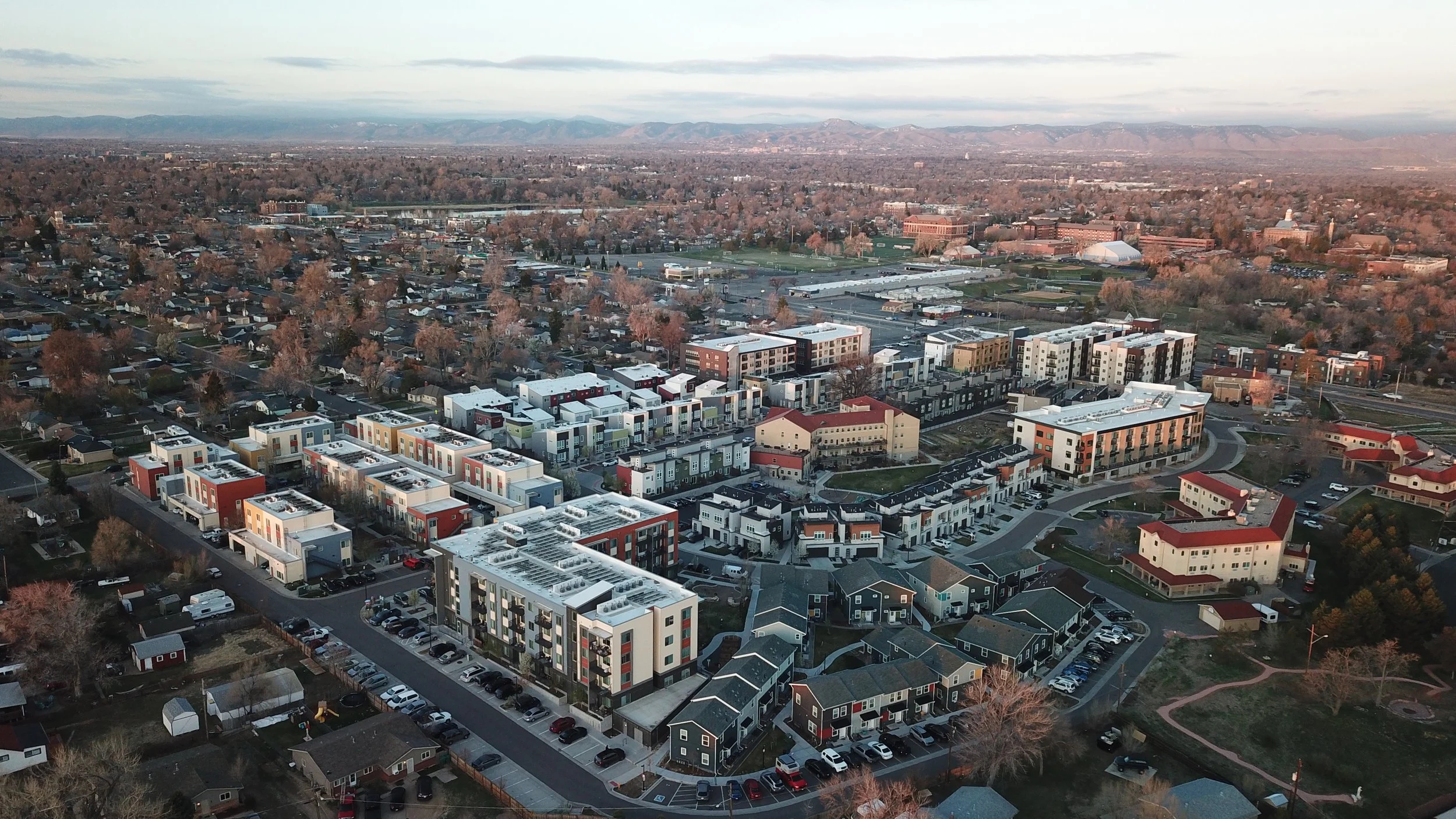 Aerial view of a suburban area with modern apartment buildings, residential houses, roads, and trees, with mountain range in the background.
