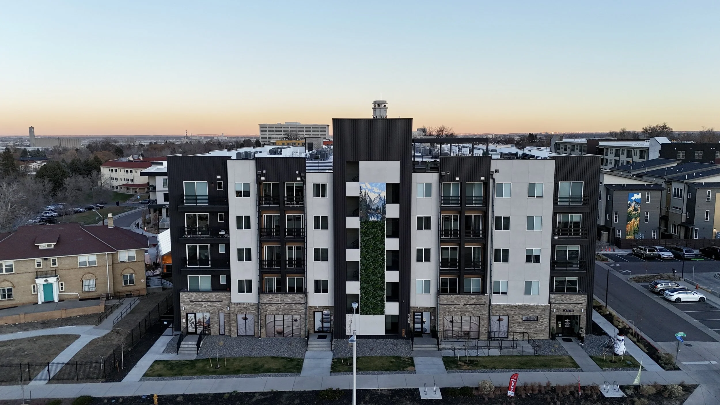 A modern multi-story apartment building with a mural of mountains and trees, located in an urban area at sunset.