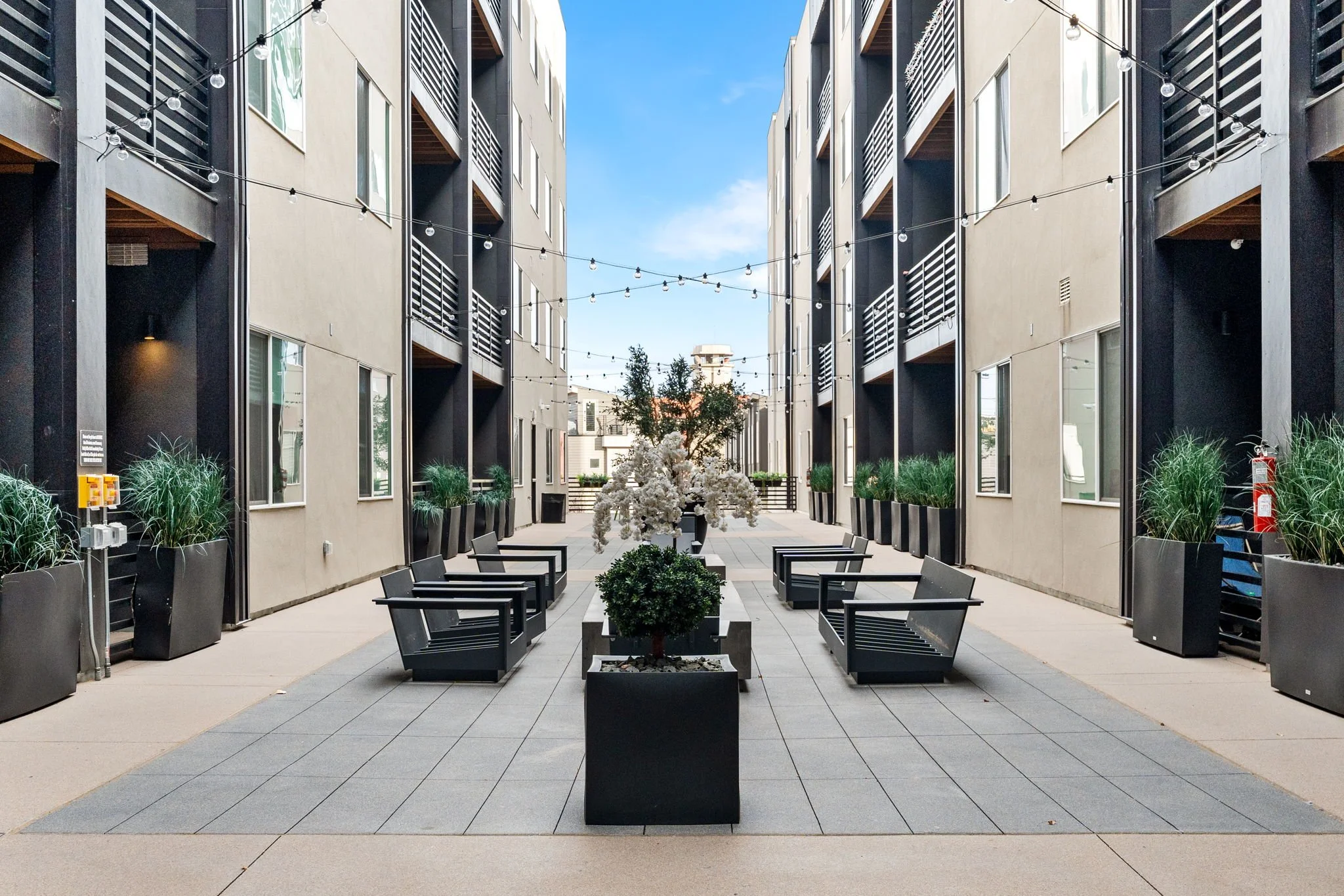 Outdoor courtyard between modern apartment buildings with string lights, potted plants, benches, and a tree in the center, under a partly cloudy sky.