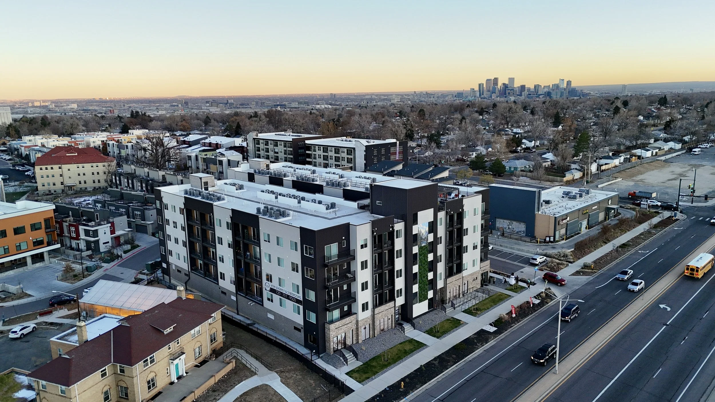 Aerial view of modern apartment complex with black and white exterior, flat roofs, and multiple air conditioning units, adjacent to a busy street with cars and a school bus, in a city with a skyline in the background.