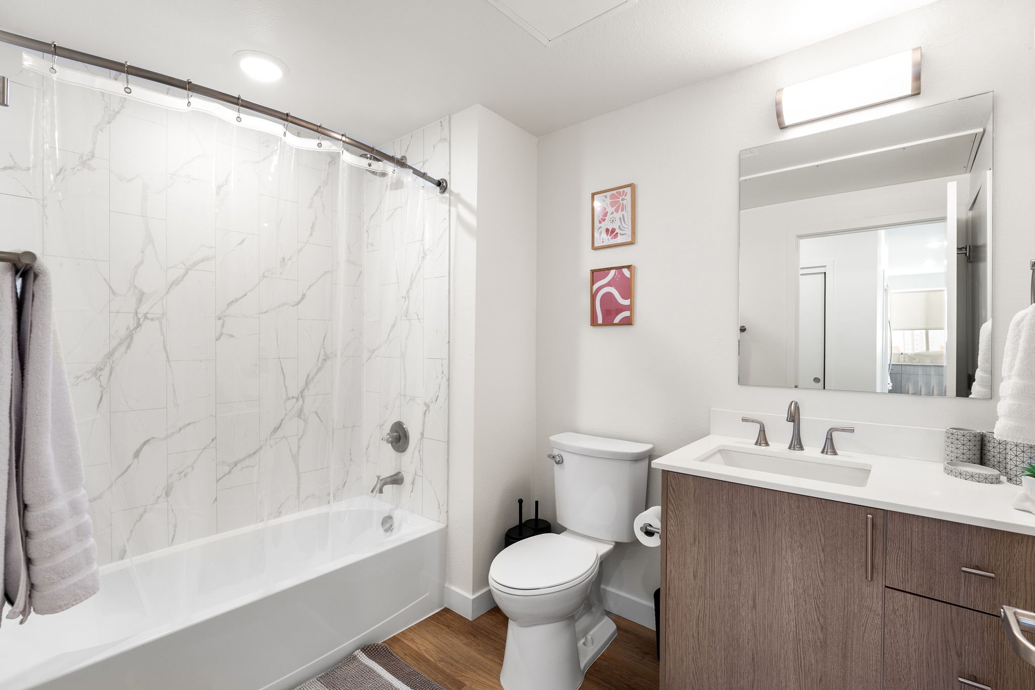 Modern bathroom with a bathtub and shower, a toilet, and a wooden vanity with a sink, mirror, towel, and artwork on the wall.