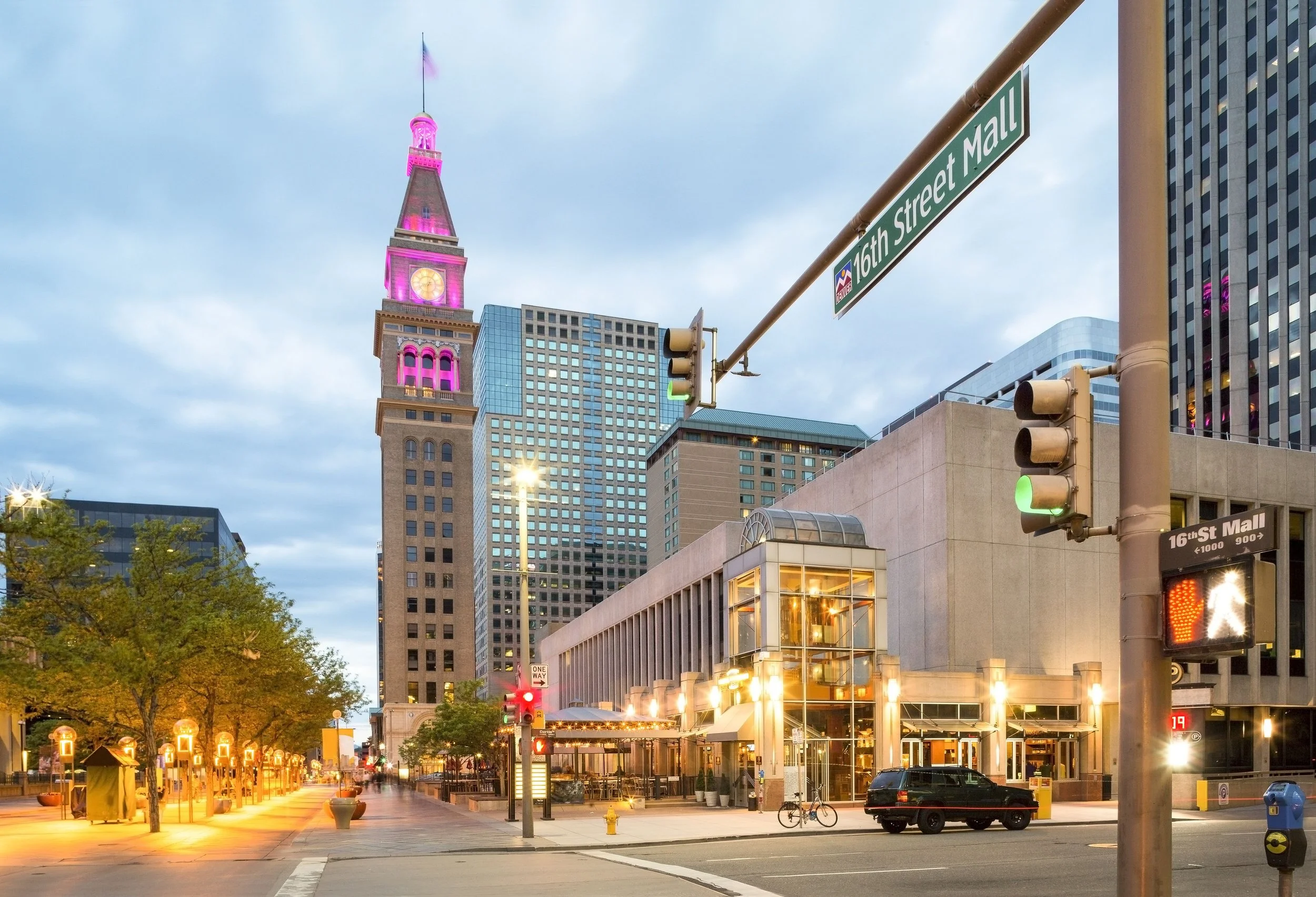 Downtown cityscape at dusk with tall buildings, green traffic lights, and a street sign indicating 16th Street Mall. Illuminated trees and street lamps line the sidewalk, and a few parked cars and bicycles are visible.