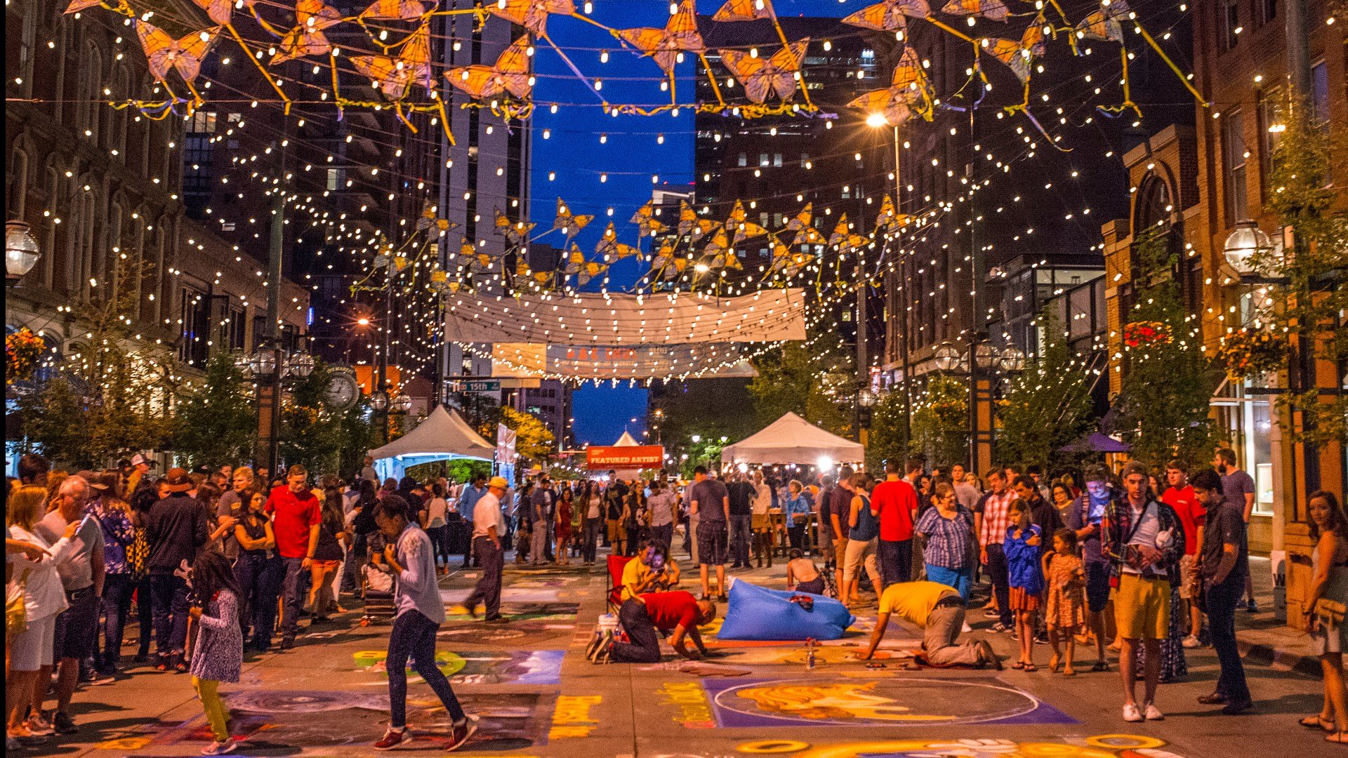 A lively street festival at night with many people gathered. The street is decorated with string lights, butterflies, and banners. Artists are painting on the ground for a chalk art event, with tents and booths in the background.