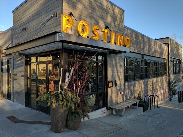 Exterior of a building with a large yellow sign that says 'POSTING' and a glass door entrance, outdoor plants, and benches.