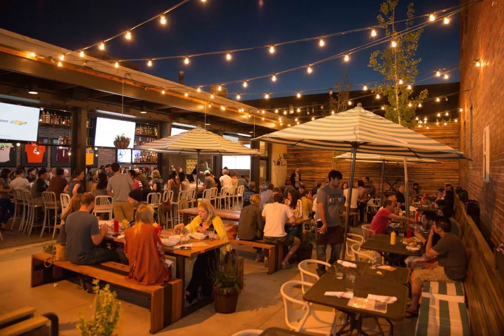 A lively outdoor restaurant or bar at night with string lights overhead, filled with people dining and socializing, some sitting at tables and others at the bar, under umbrellas and against a brick wall.