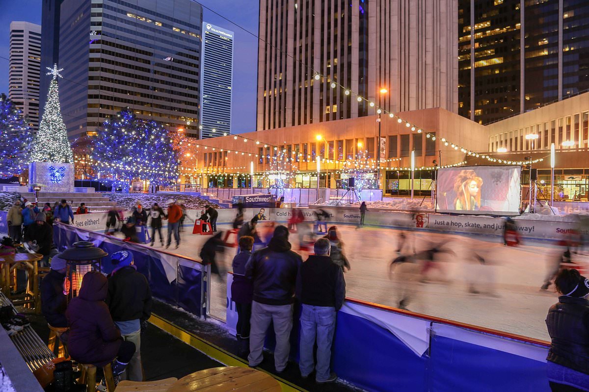 People ice skating at an outdoor rink in a city square decorated for Christmas with blue and white lights, a Christmas tree, and snow fall at dusk.