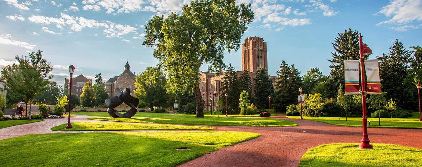 A well-maintained park on a bright, sunny day with a paved red brick pathway, lush green grass, mature trees, a modern sculpture, and historic buildings in the background surrounding the University of Denver.