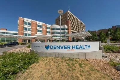 Denver Health hospital with a large sign in front displaying the name, surrounded by grass and rocks, with a modern multi-story building in the background under a clear blue sky.