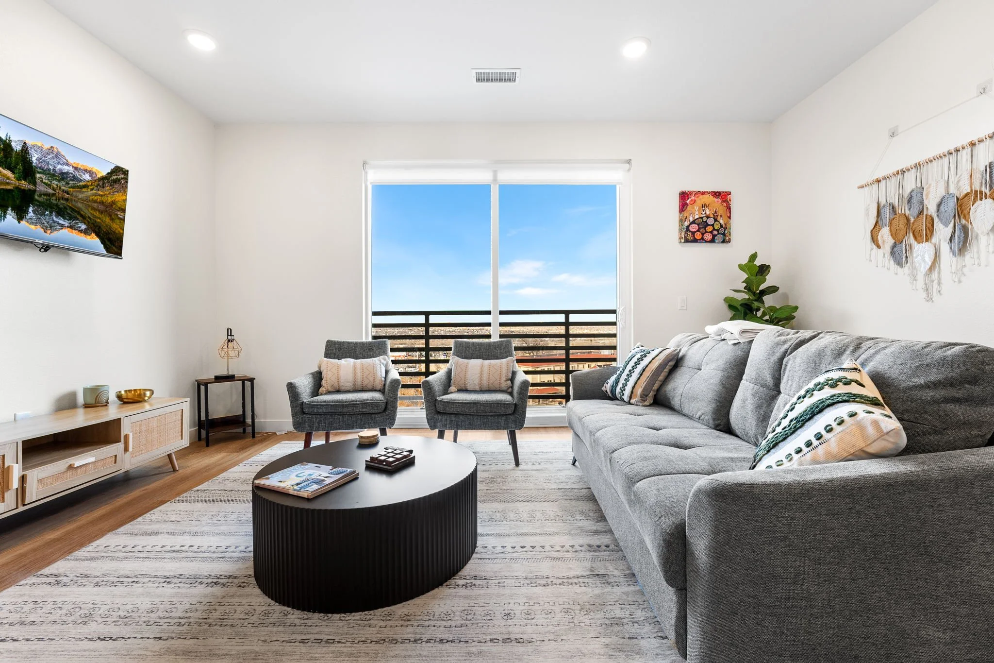 Modern living room with a large gray sofa, two matching armchairs, a black coffee table, a white media console, wall-mounted TV, sliding glass door with view of cityscape, patterned rug, and decorative wall hangings.
