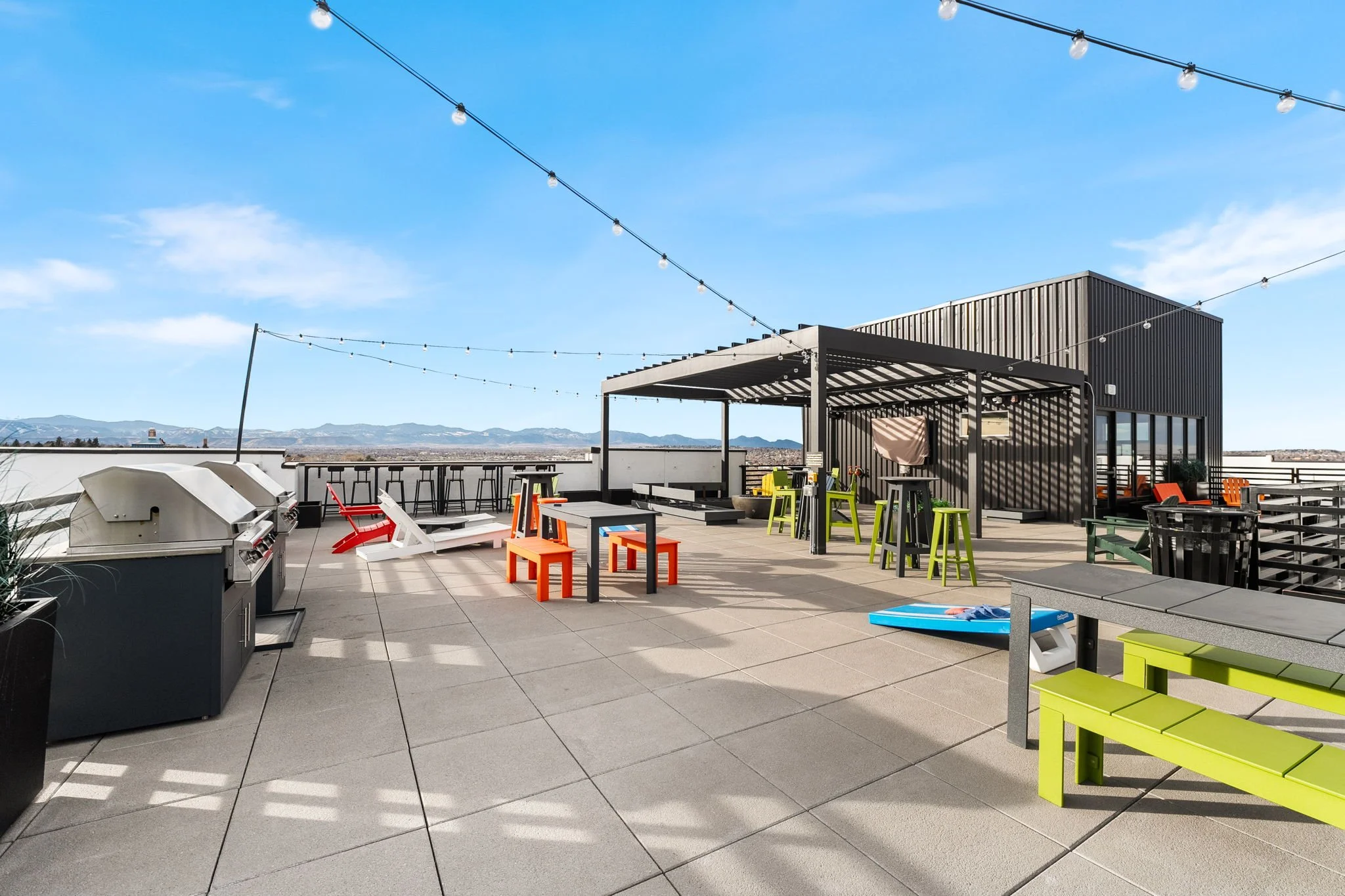 Rooftop outdoor patio with string lights, colorful furniture, grills, and a city view in the background.
