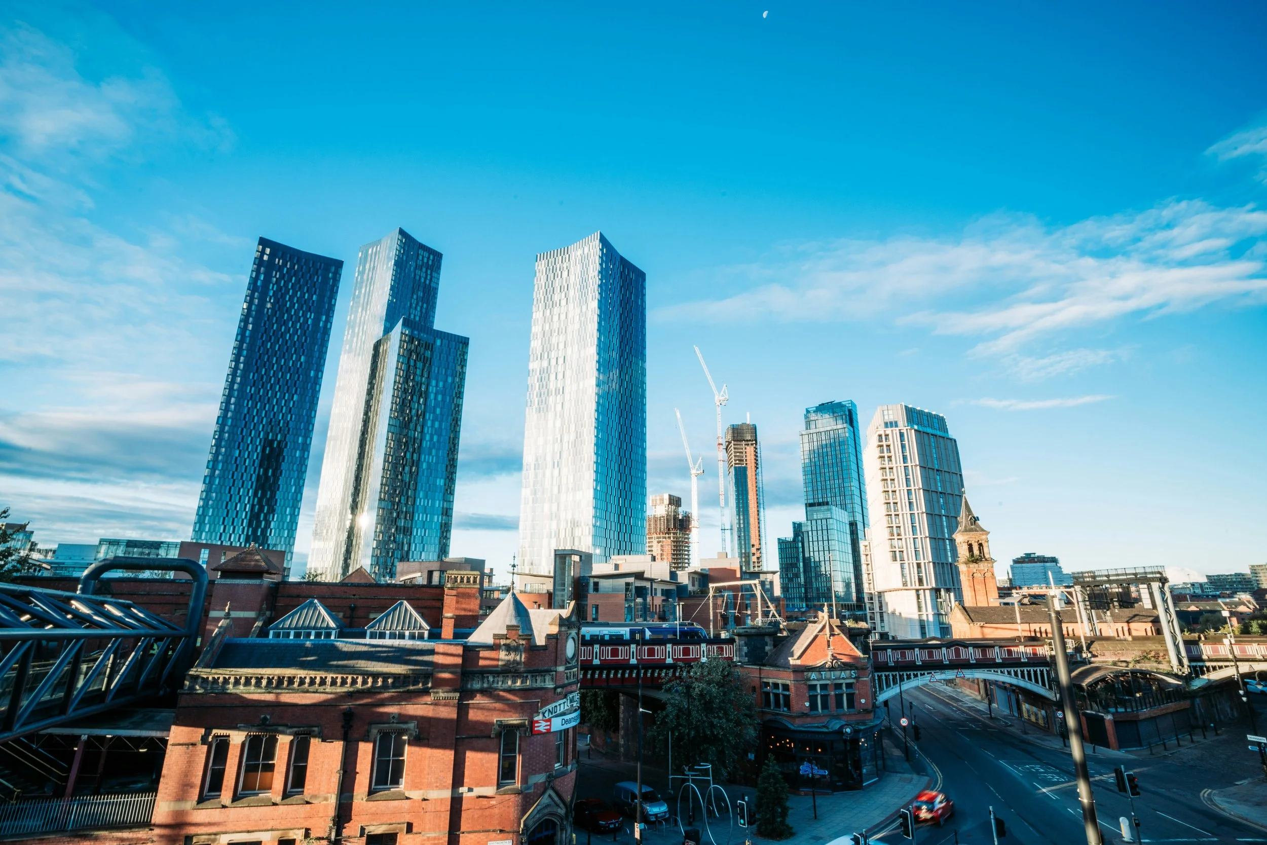 Cityscape with tall modern skyscrapers during daytime under blue sky with some clouds and a visible moon, with older brick buildings and a street with cars in the foreground.
