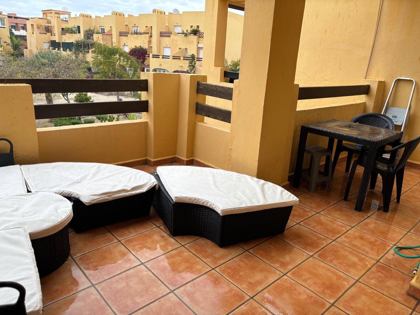 Balcony with wicker furniture, including a cushioned lounge chair and a small table, overlooking a neighborhood of yellow buildings with trees and rooftops.
