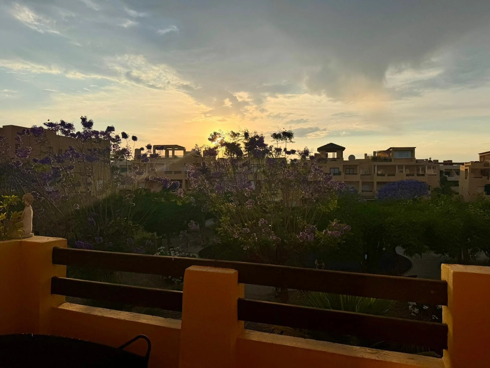 Sunset over a residential area with trees and flowering bushes, viewed from a balcony with a yellow wall and wooden railing.