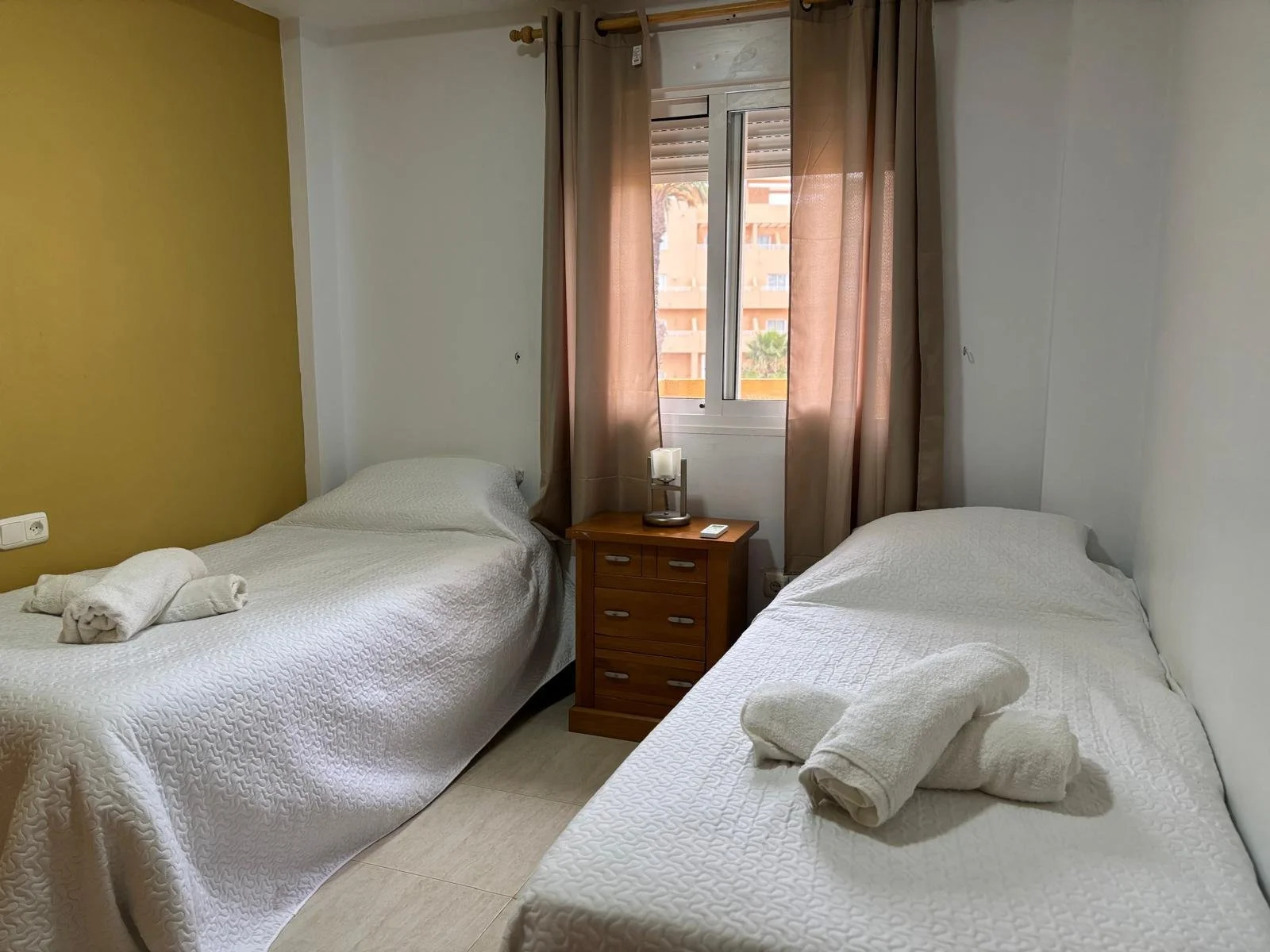 A hotel room with two single beds covered in white quilted bedspreads, each with rolled towels on top. There is a wooden nightstand between the beds with a small lamp, a remote control, and a puzzle piece on it. The room has a window with beige curtains, and outside buildings are visible.