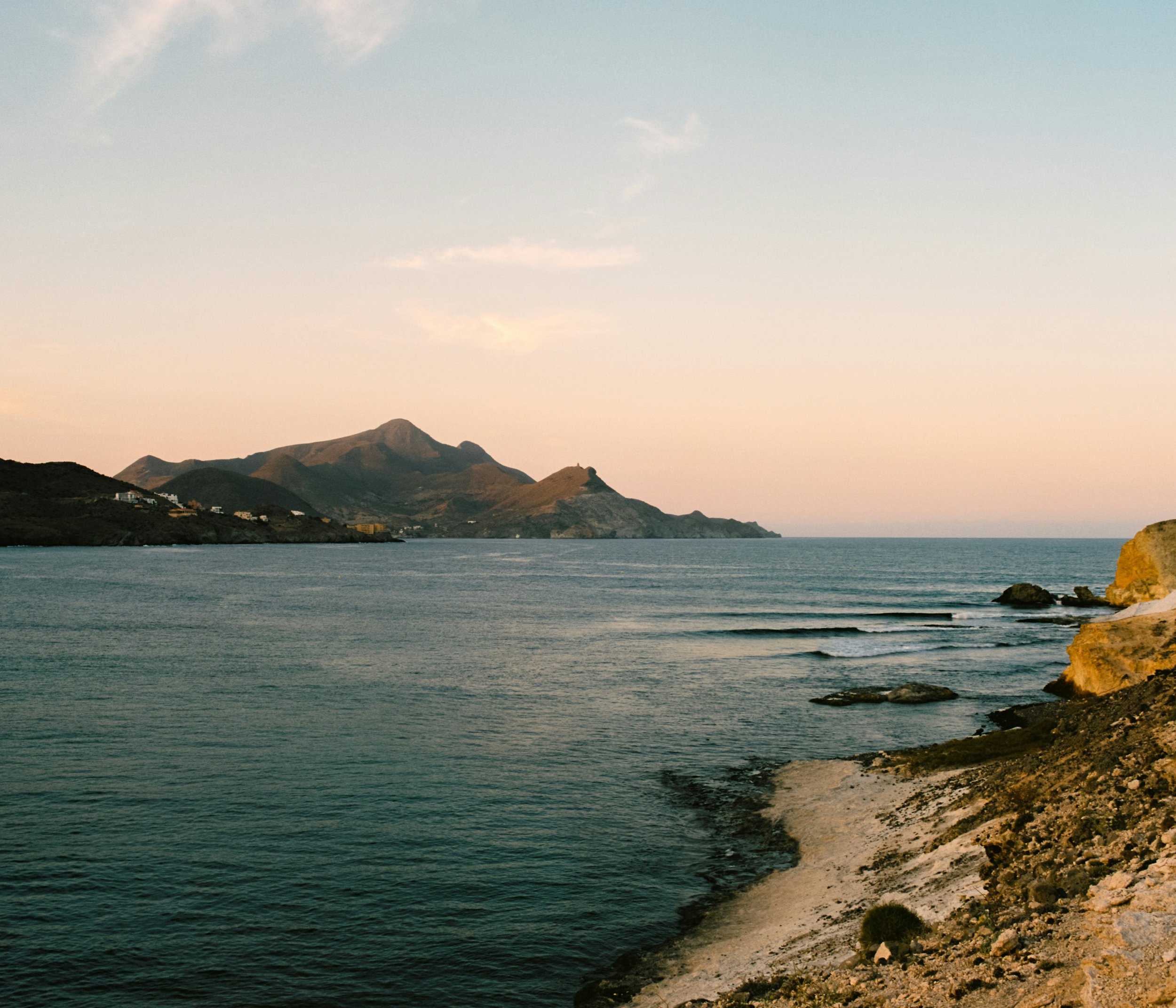 A coastal landscape at sunset with mountains in the distance, calm sea, and rocky shoreline in the foreground.