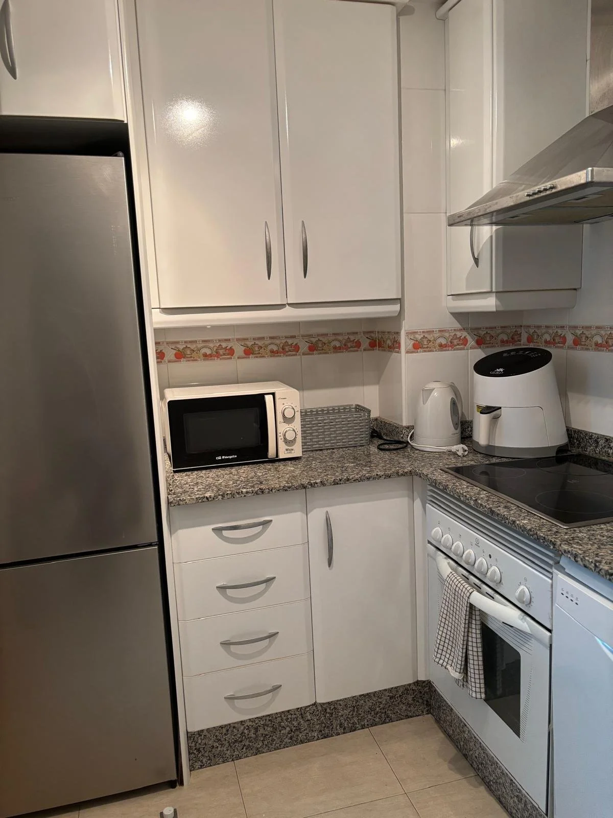 Small kitchen with white cabinets, a stainless steel refrigerator, a microwave, an electric kettle, a toaster oven, and an electric stove with oven. Granite countertops and tiled backsplash with decorative border.