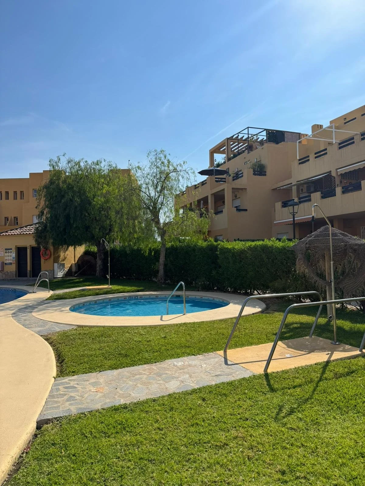 A swimming pool and a hot tub in a residential complex with green grass, trees, and bushes under a clear blue sky.