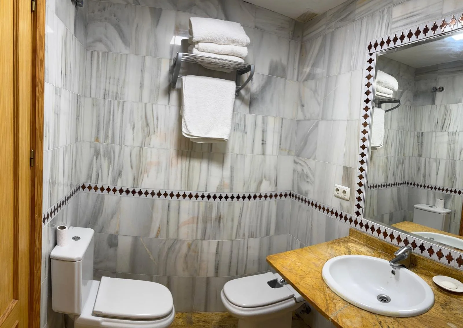 Small bathroom with marble wall tiles, a toilet, a mirror with decorative border, a sink with a yellowish-brown countertop, and a towel rack with three towels.