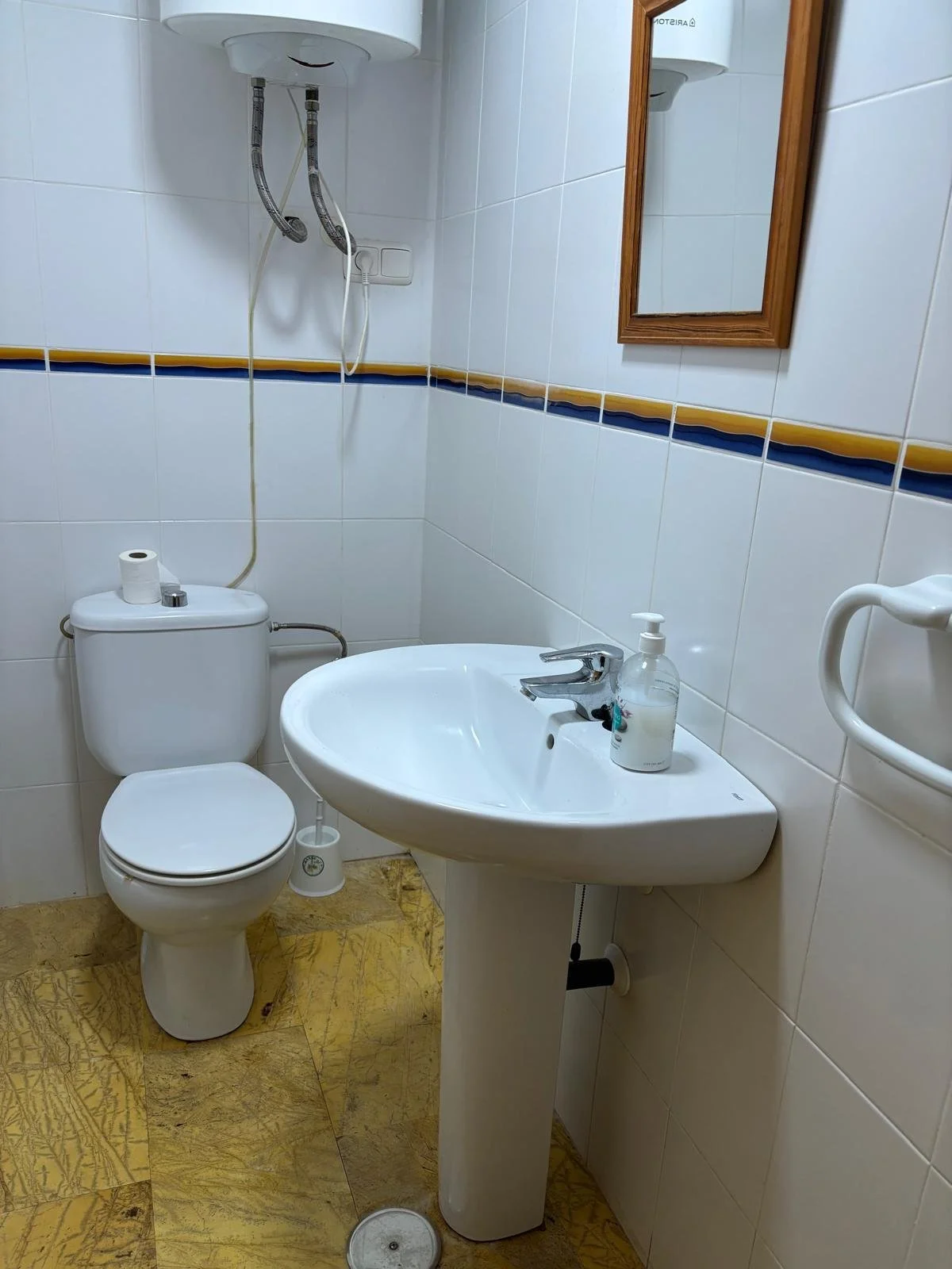 Small bathroom with white tiled walls and a yellow patterned floor, featuring a toilet, a wall-mounted sink with a bottle of hand soap, a mirror, a water heater, and a towel holder.