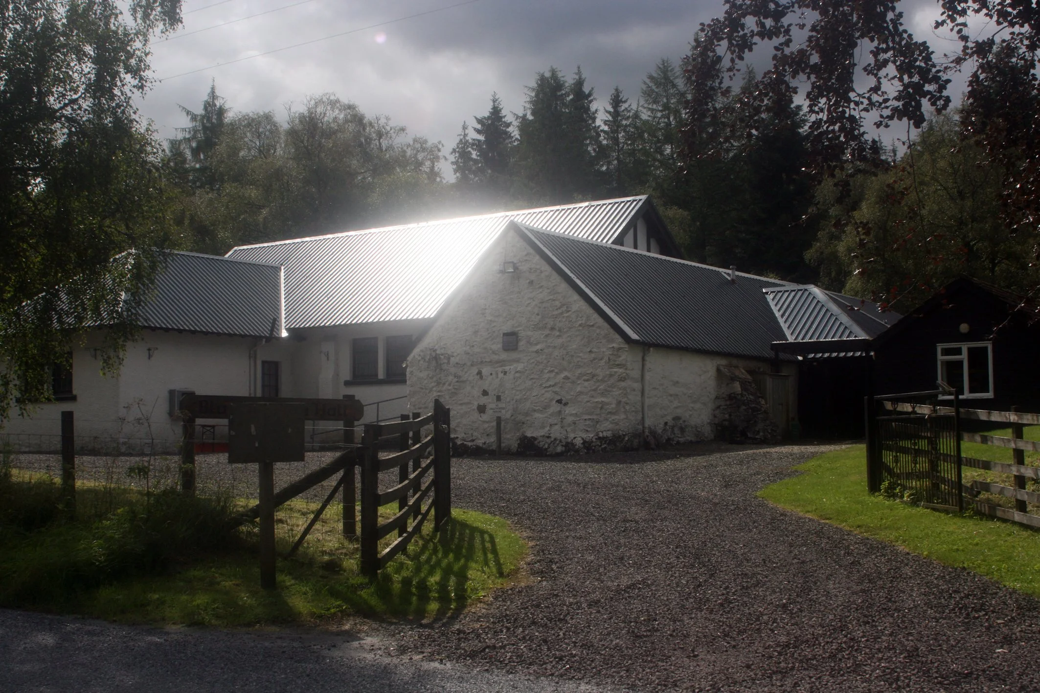 Hall with white walls and dark metal roof, surrounded by trees and a wooden fence, with cloudy sky overhead.