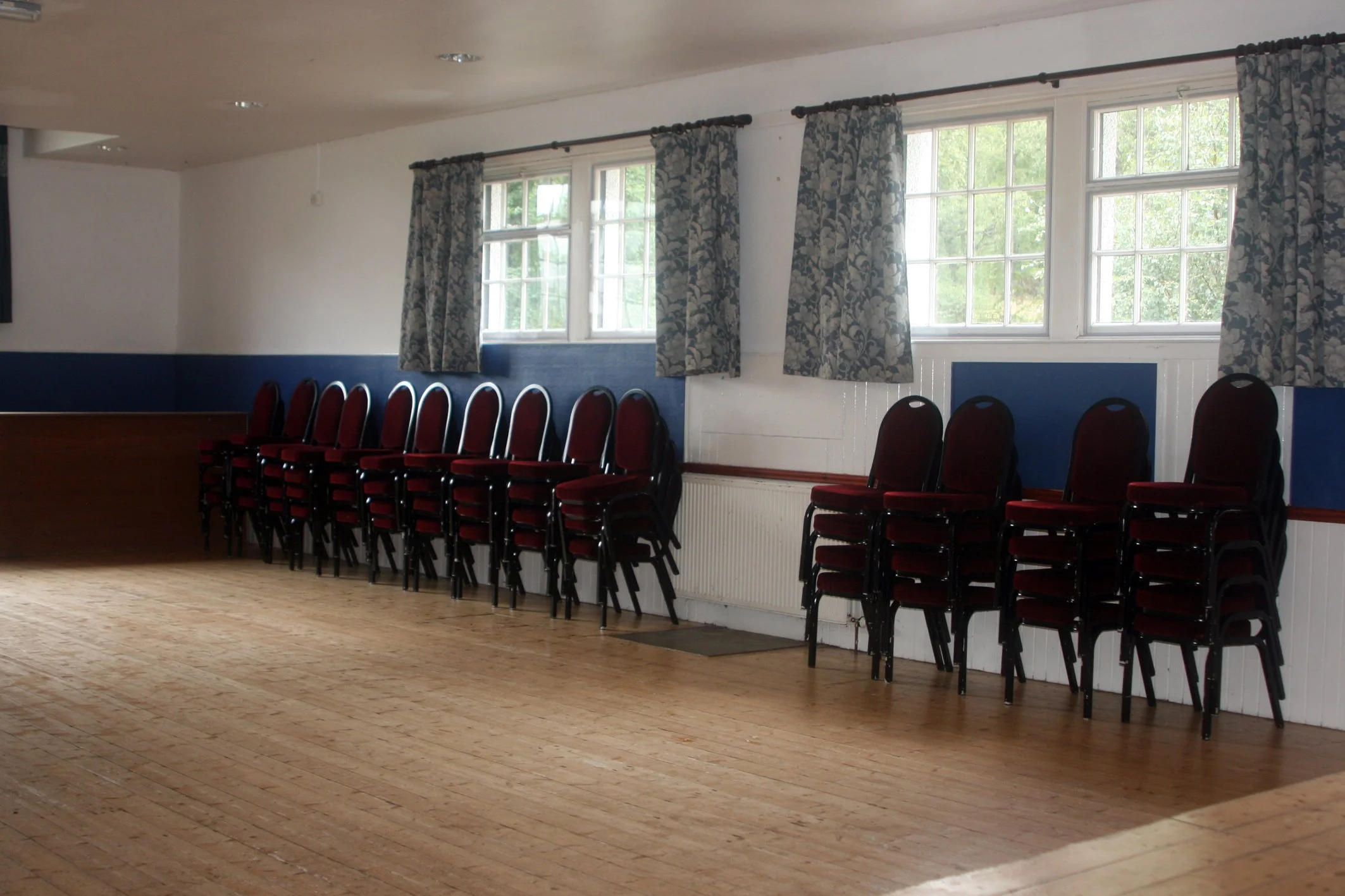 An empty conference or banquet room with a row of red and black chairs along the wall, large windows with floral curtains, wood flooring, and white walls with a blue accent.