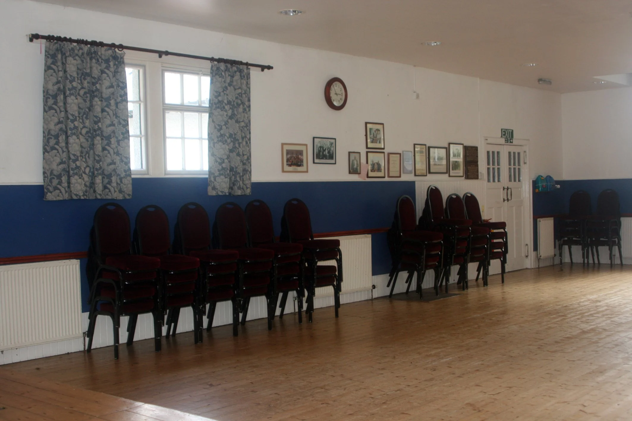 Empty room with a row of stacked chairs along the wall, windows with floral curtains, framed pictures on the wall, a clock, and an exit sign above a door.
